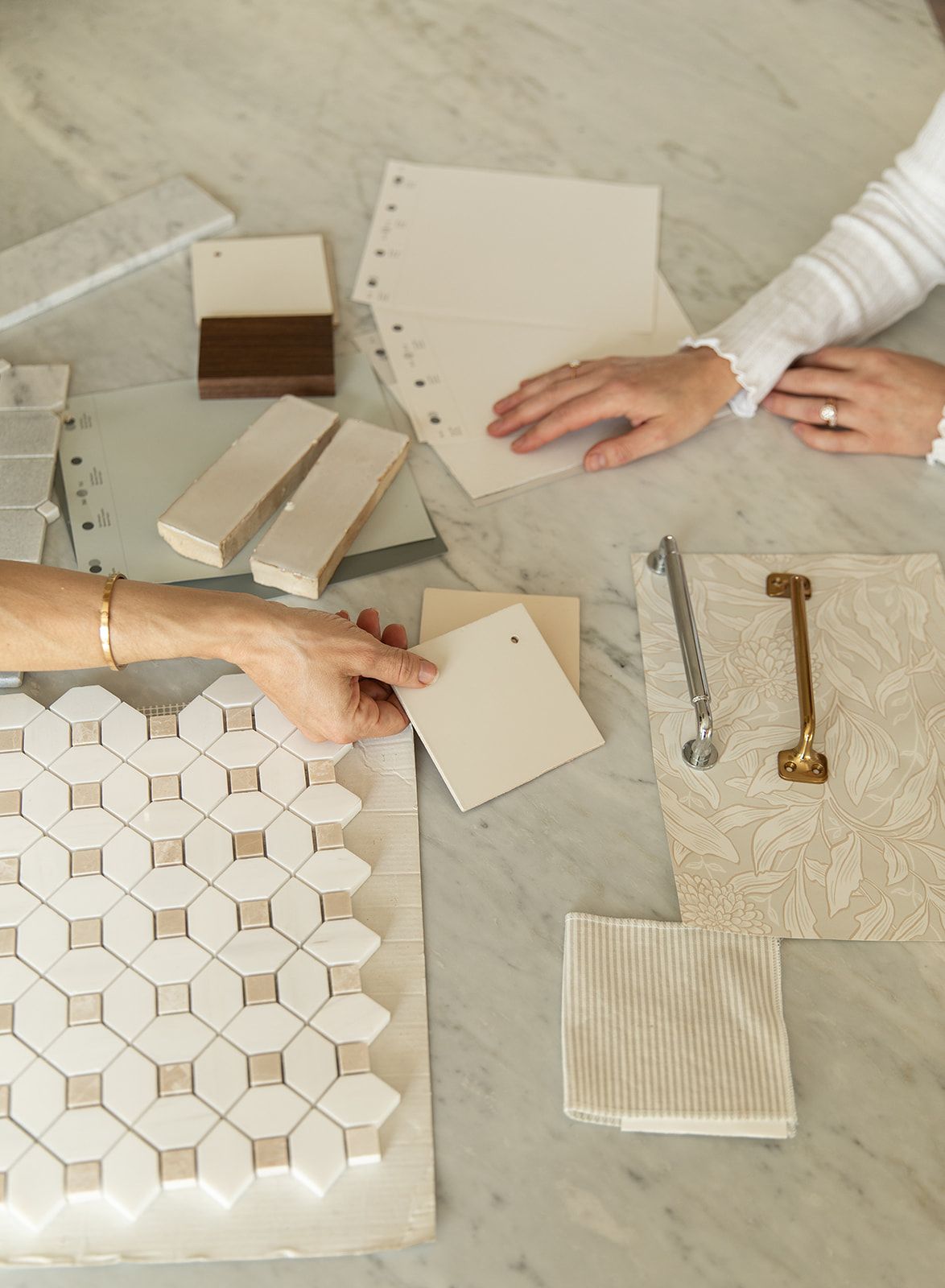 Two designers review tile samples, paint swatches, and hardware finishes on a marble table during an interior design consultation.