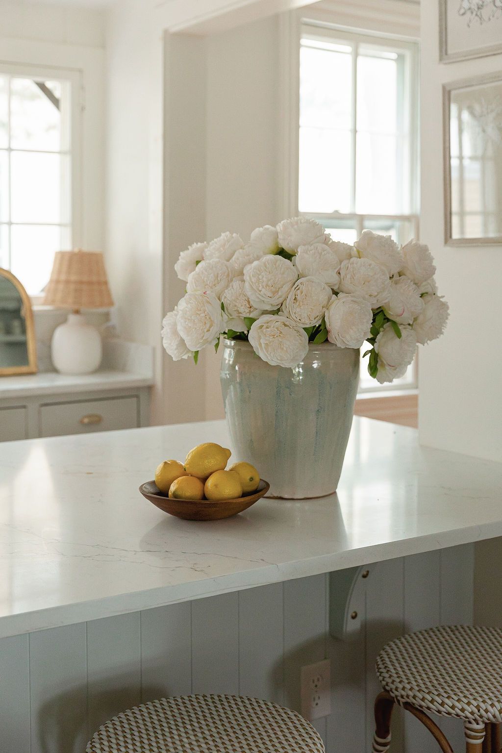 White peonies in ceramic vase with fresh lemons on marble counter in bright kitchen interior.