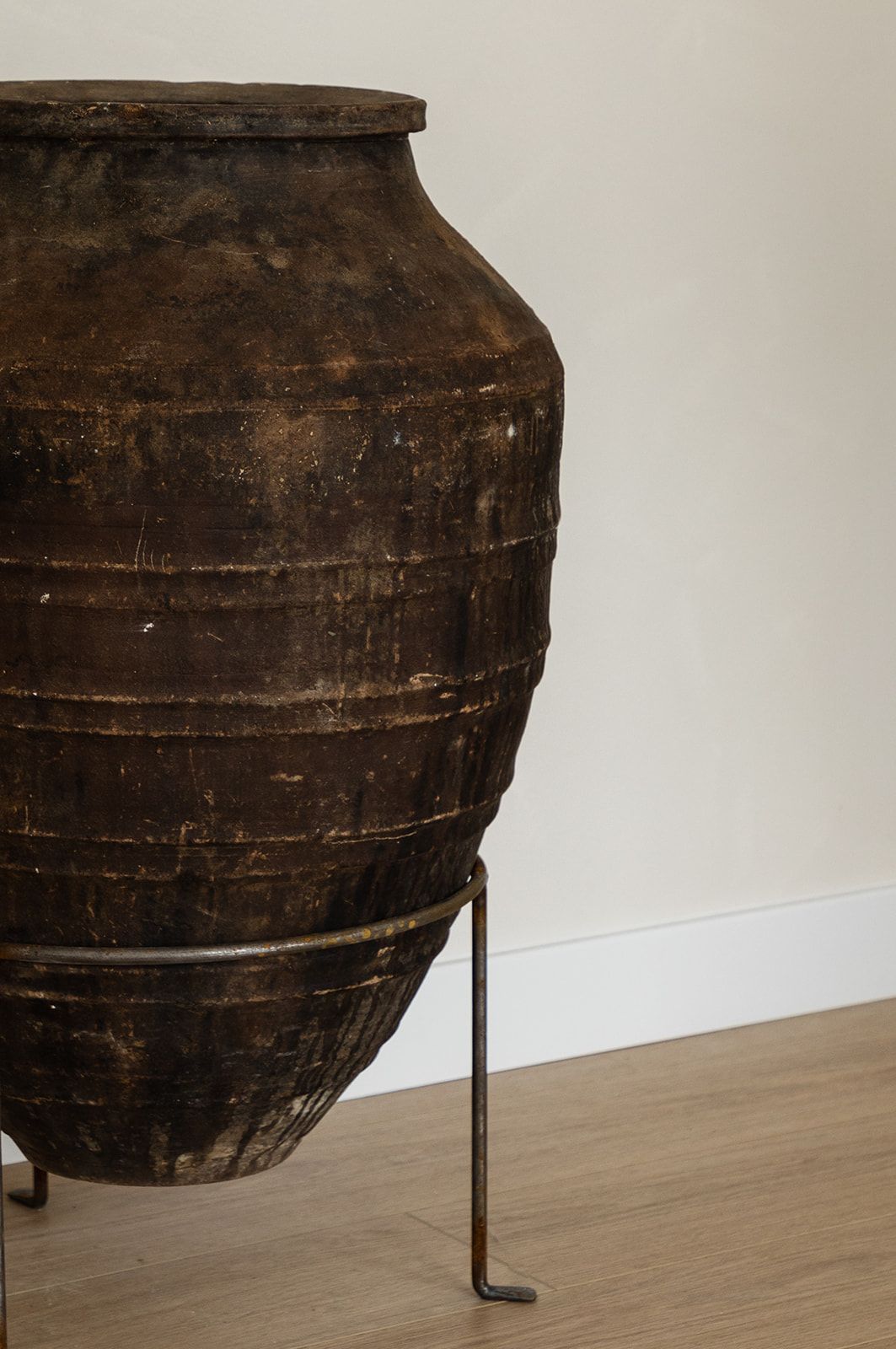 Large antique earthenware vessel on metal stand against white wall and wooden floor.