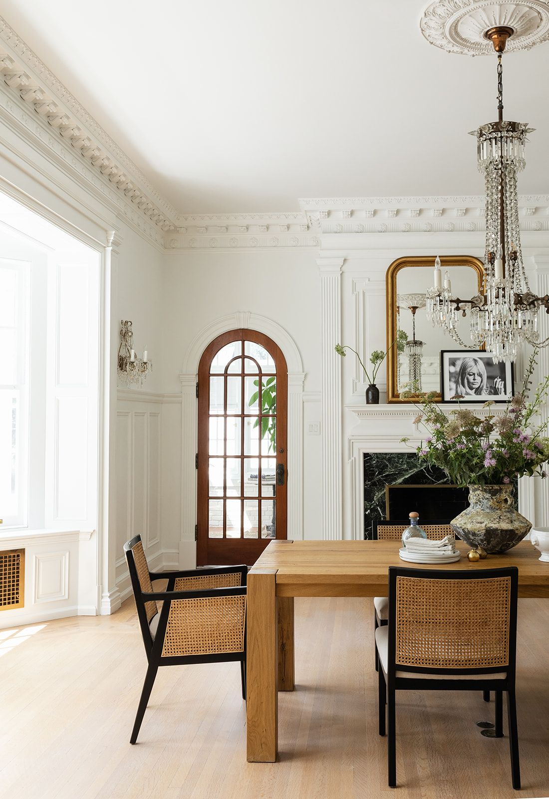 Modern dining area featuring arched wooden door, gold mirror above fireplace, and cane-backed chairs.