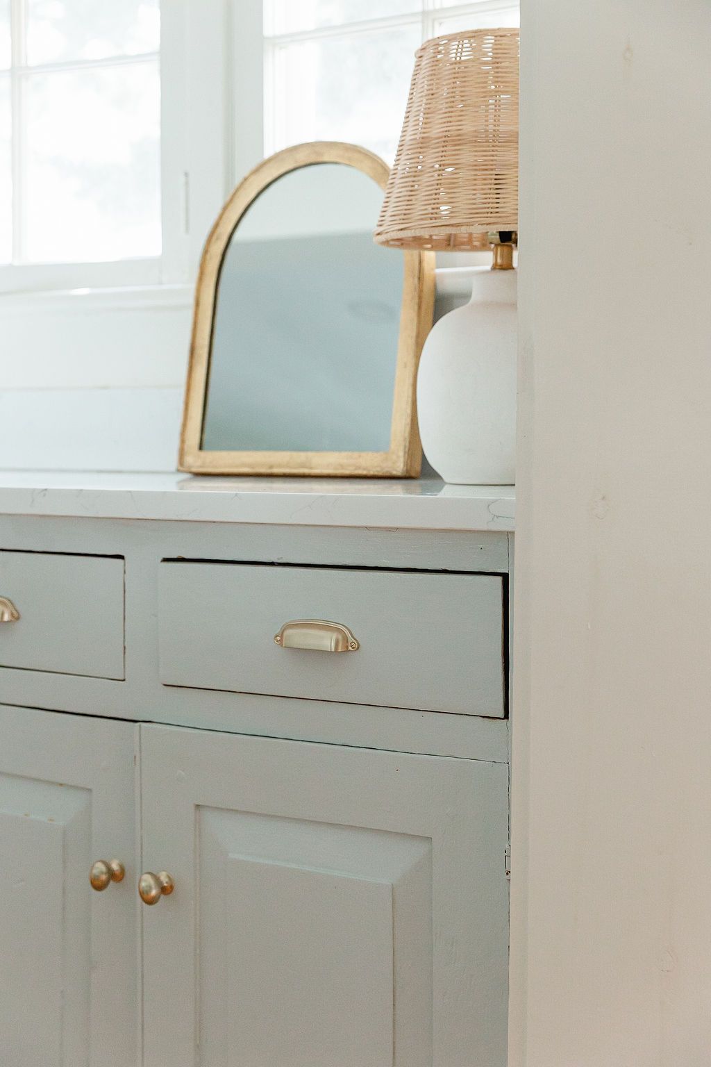 Sage green cabinet with brass hardware and arched mirror on top with rattan lamp shade.
