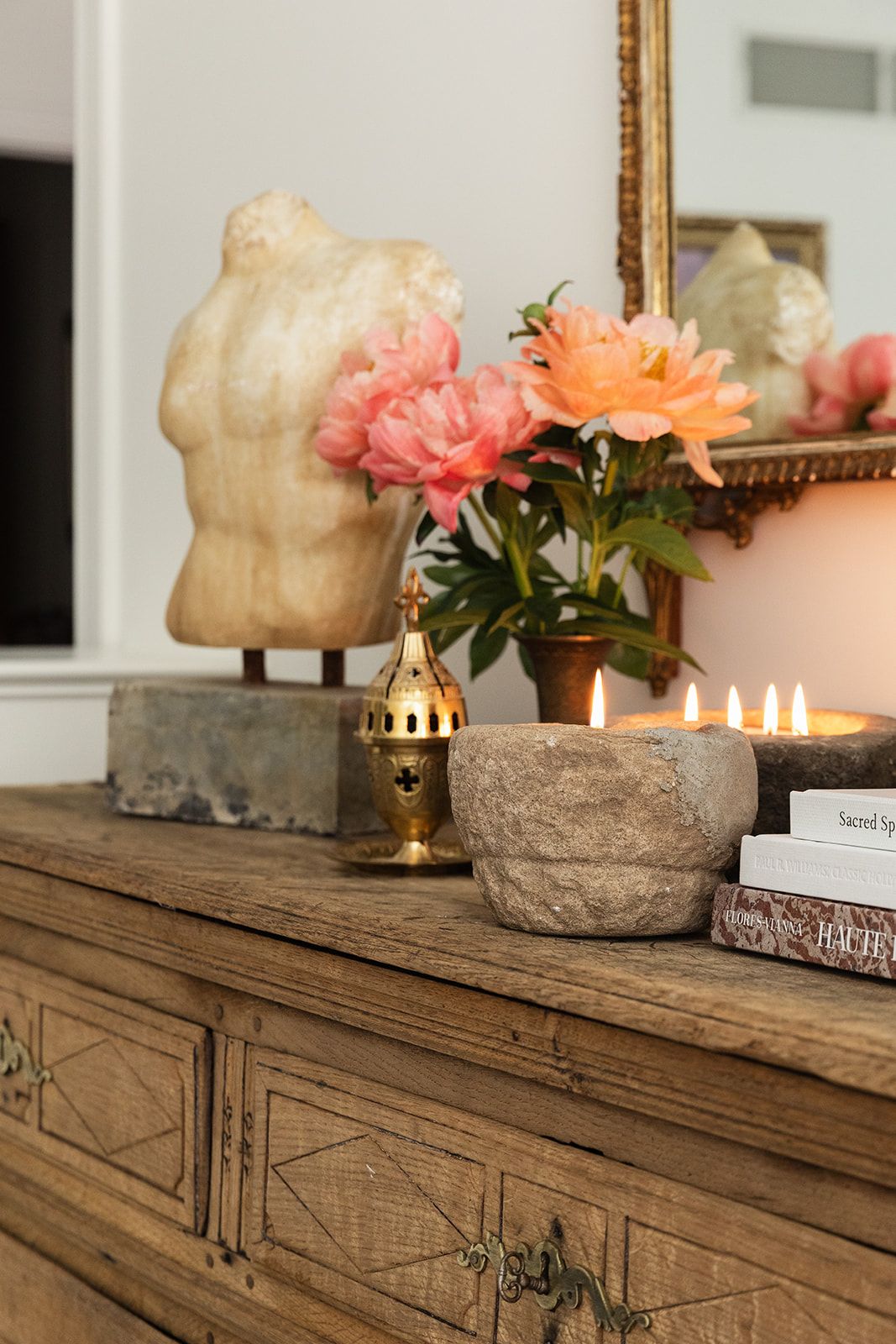 Rustic wooden dresser styled with a torso sculpture, pink peonies, gold lantern, stone candle bowl, and stacked books.