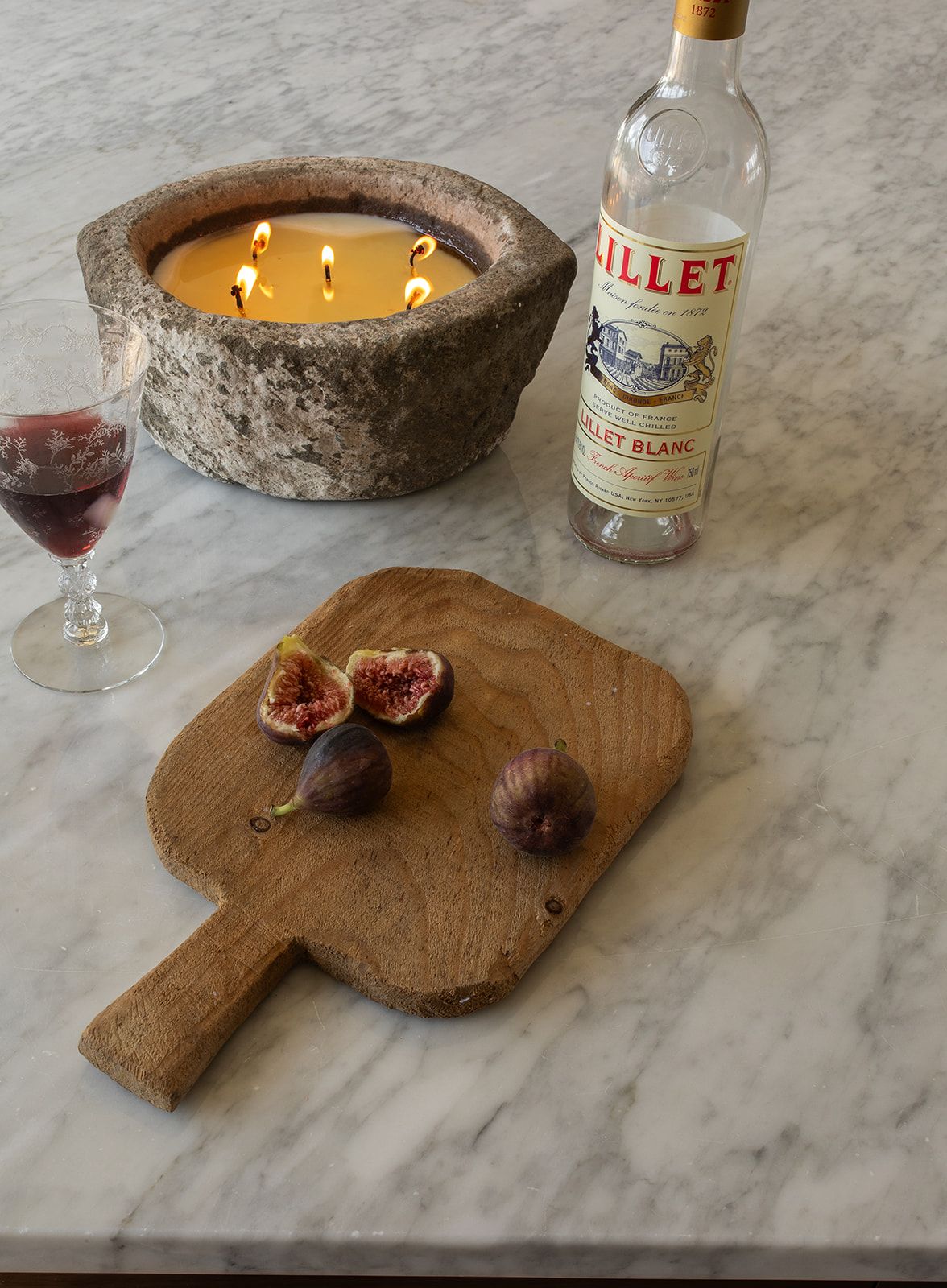 Rustic marble surface with figs on wood board, Lillet Blanc bottle, red wine glass, and stone candle holder.