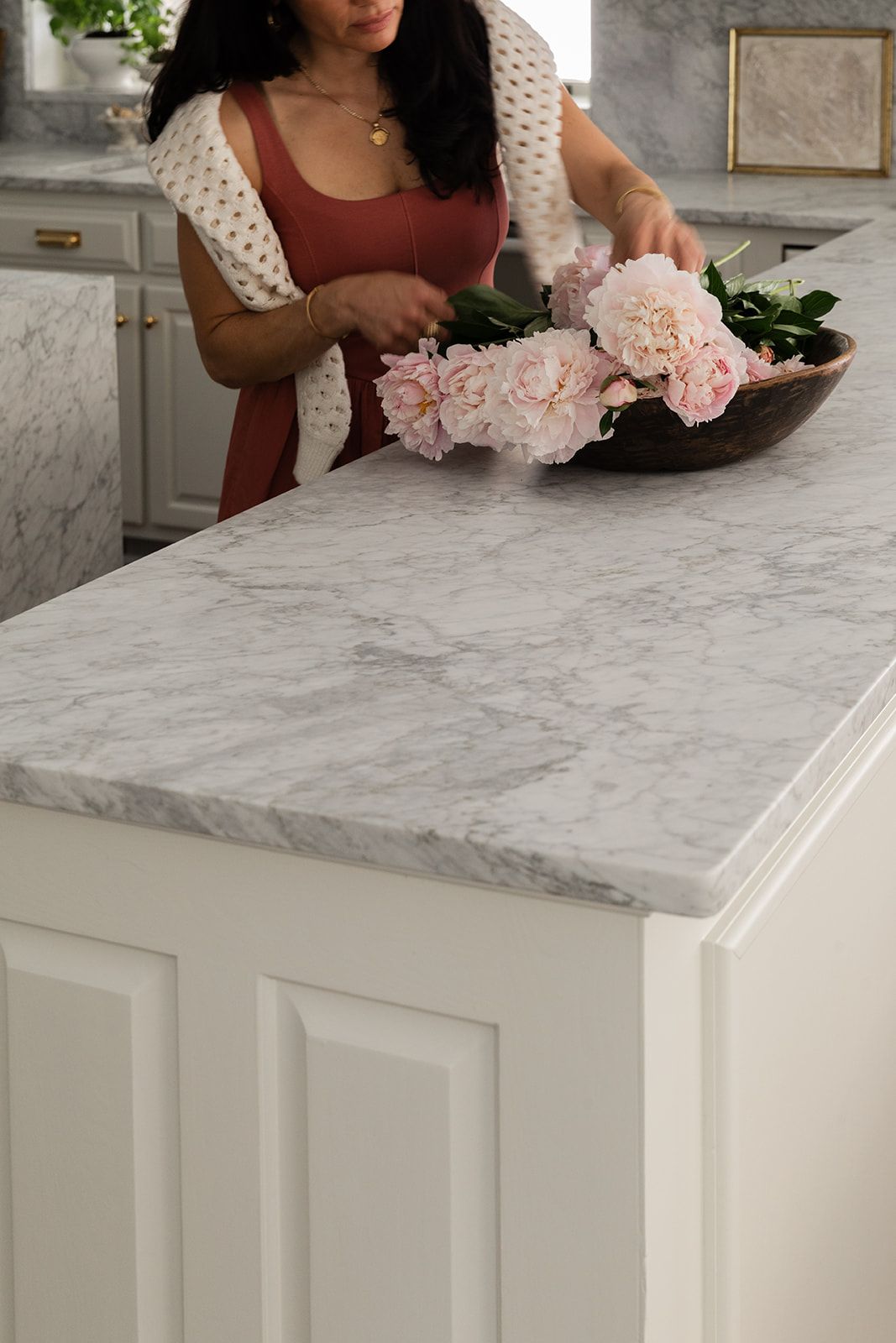 Woman arranging pink peonies in a wooden bowl on a white marble kitchen island countertop.
