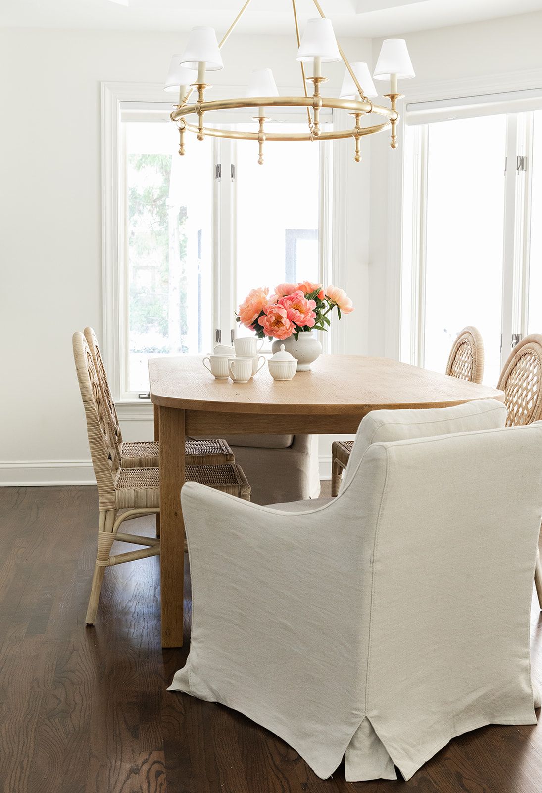 Bright dining room with oval wood table, rattan chairs, white slipcovered chair, gold chandelier, and pink floral centerpiece.
