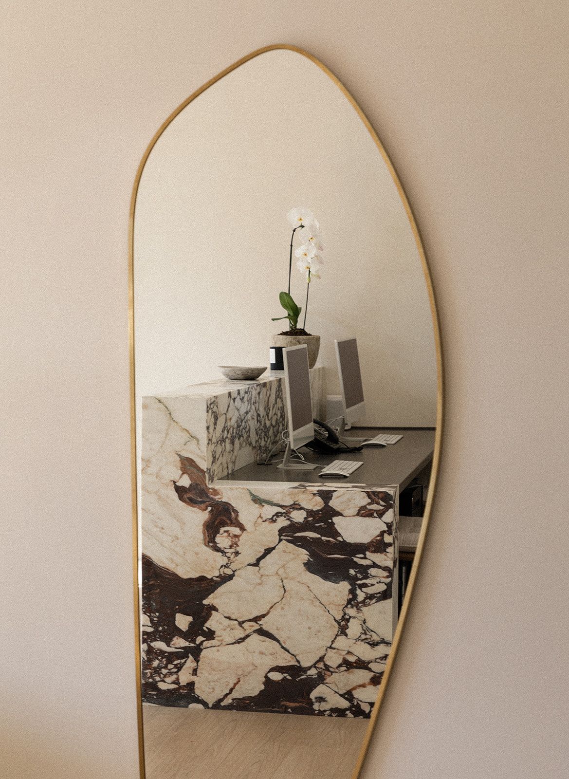 A minimalist gold-framed mirror on beige wall reflects a white orchid plant and abstract black and white marble pattern below.