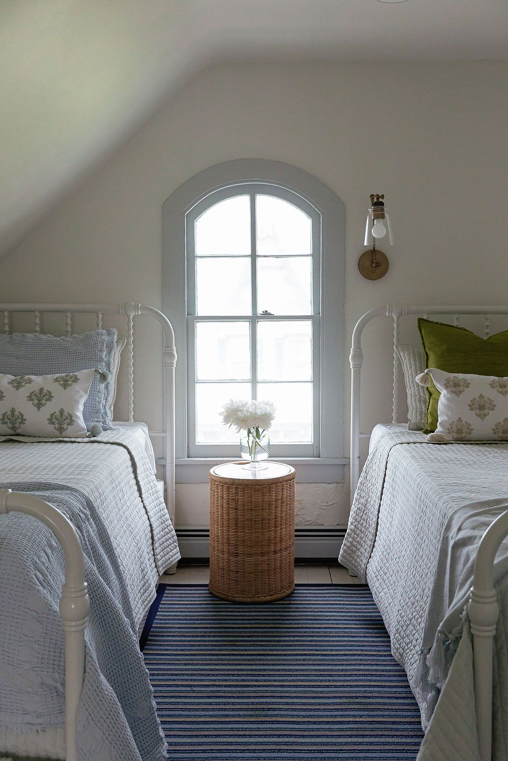 Twin white iron beds flank a wicker side table beneath an arched window in a bright attic bedroom with striped carpet.