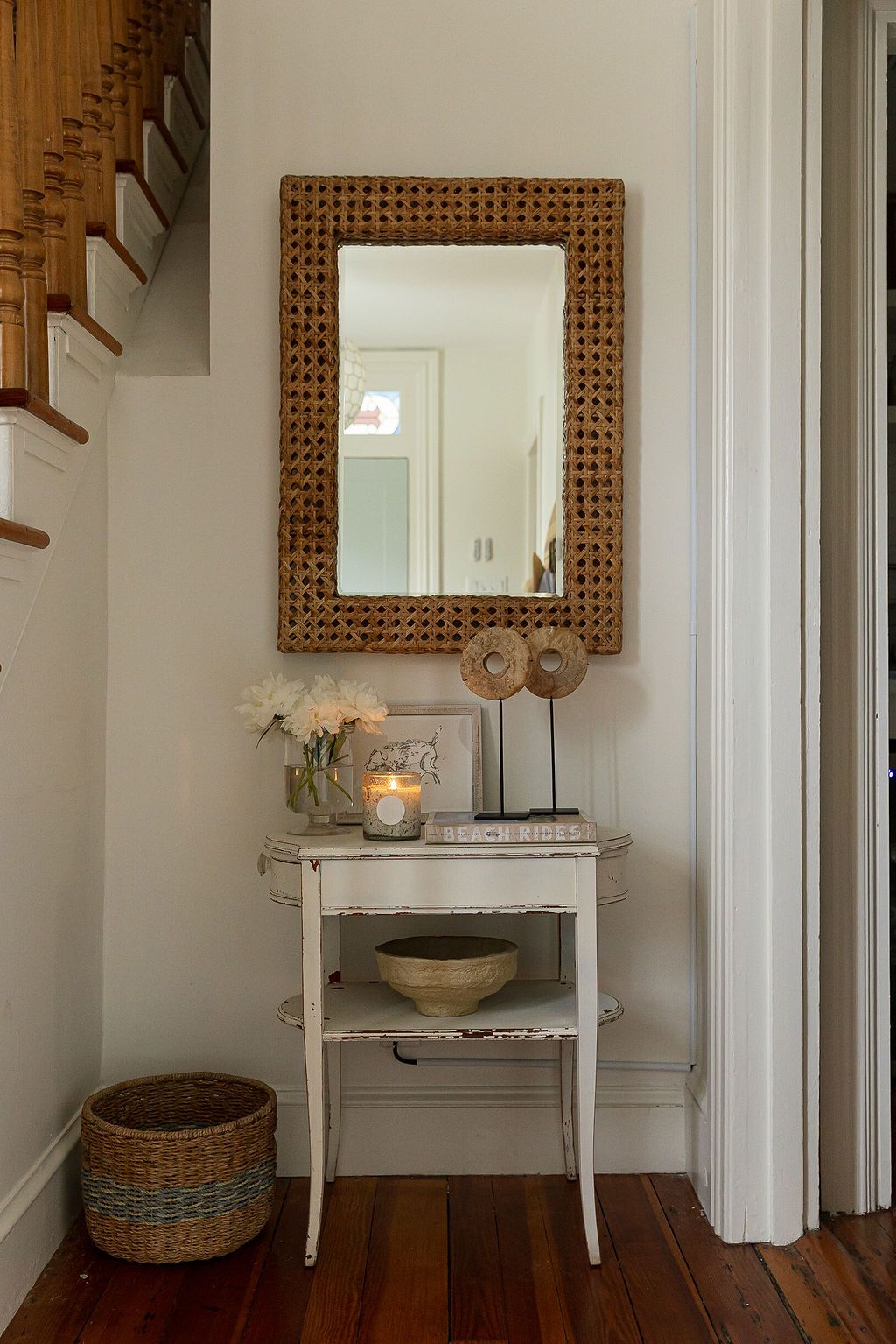 A vintage white console table with woven mirror and decorative accents sits in a cozy nook beneath wooden stairs.