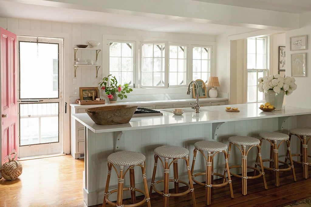 Bright modern farmhouse kitchen with white walls, wooden barstool seating, and natural light streaming through multiple windows.