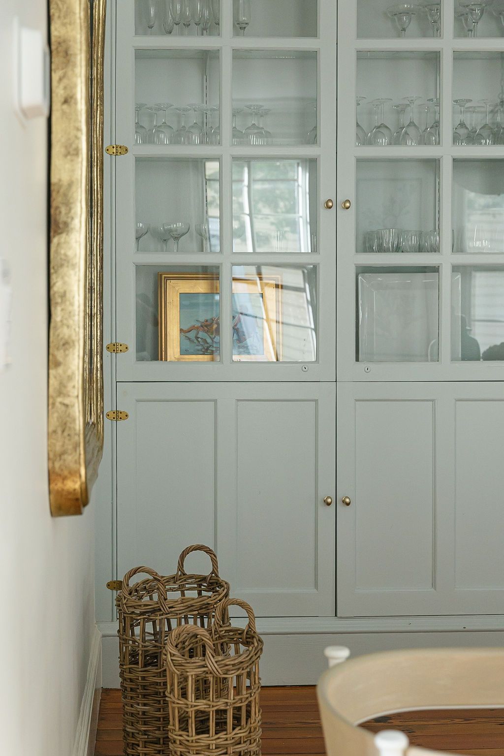 Light blue built-in cabinet with glass doors displaying glassware, with woven baskets on the floor.