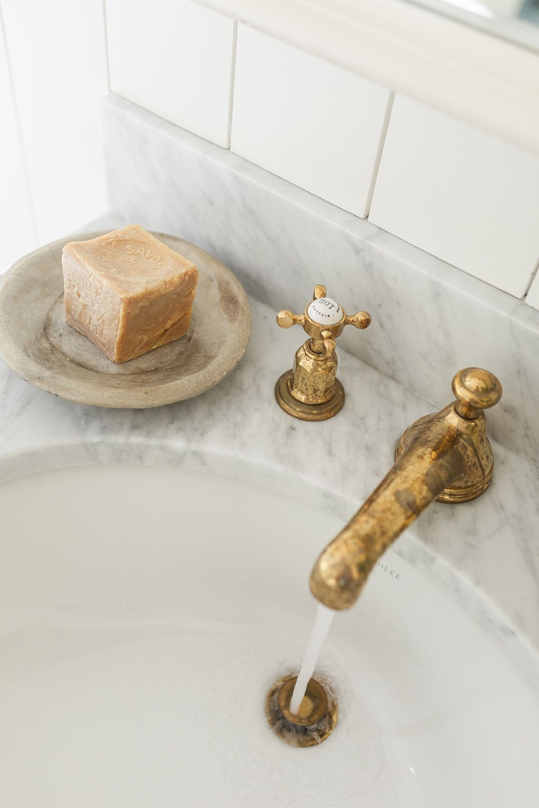 Vintage brass faucet running water into white sink with marble countertop and natural soap bar on stone dish.