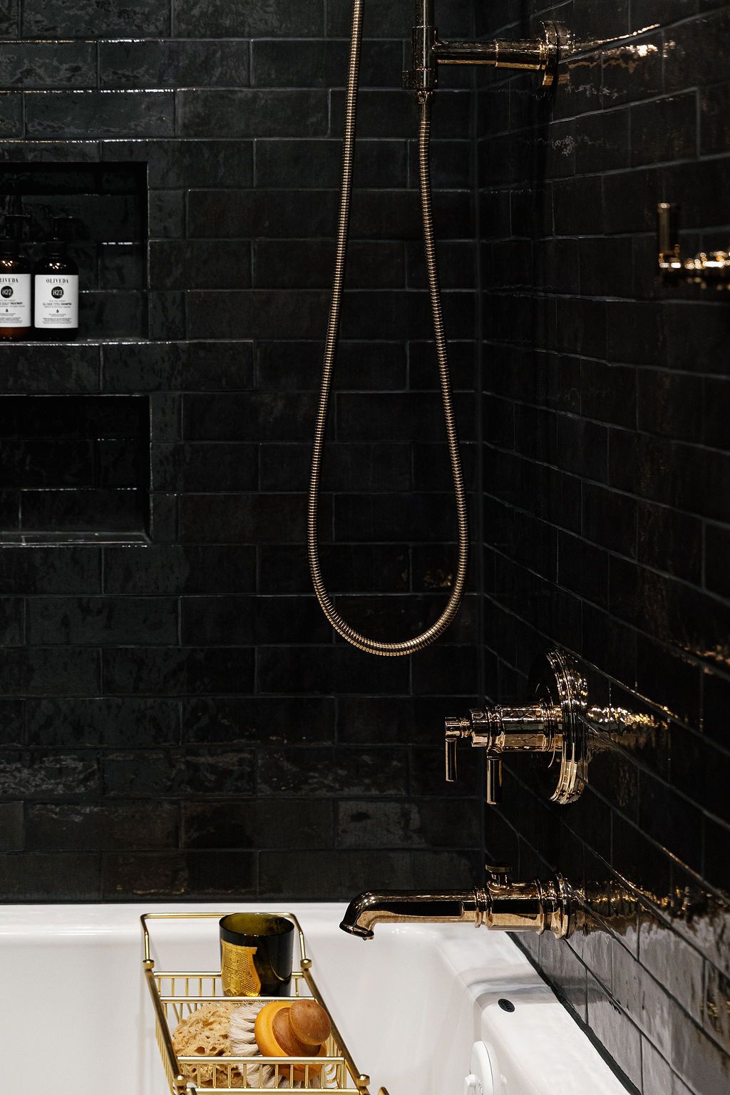 Dark tiled bathroom interior with gold fixtures and freestanding wooden storage rack holding accessories and towels.