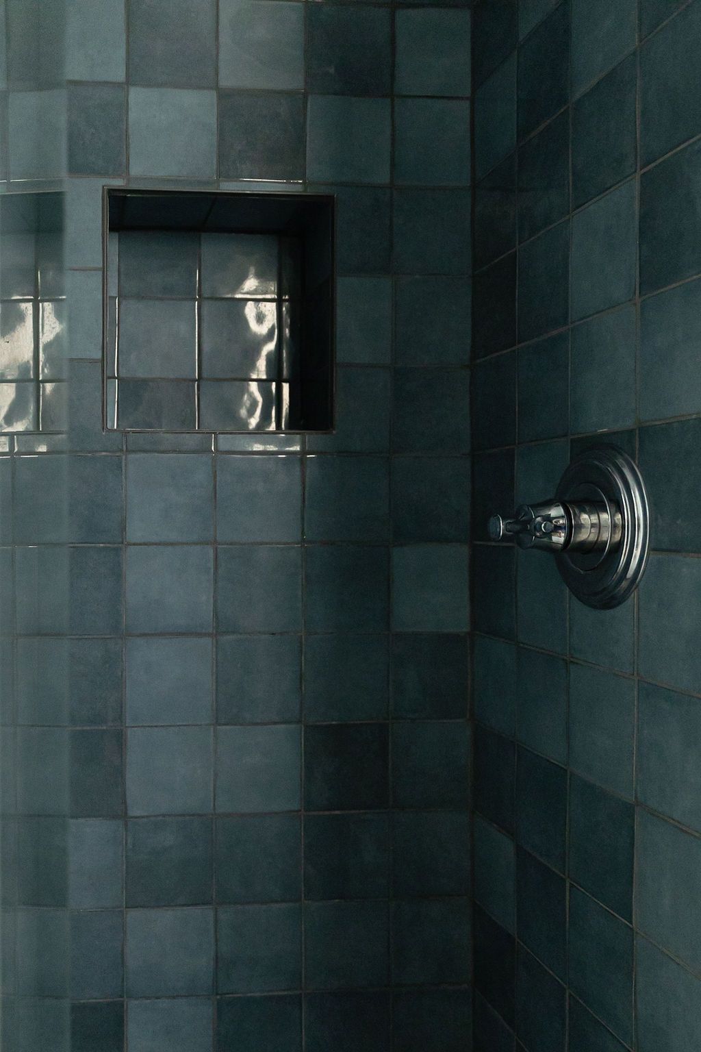 A modern bathroom shower with dark teal tile walls, a chrome showerhead, and built-in glass shelf niche illuminated by light.