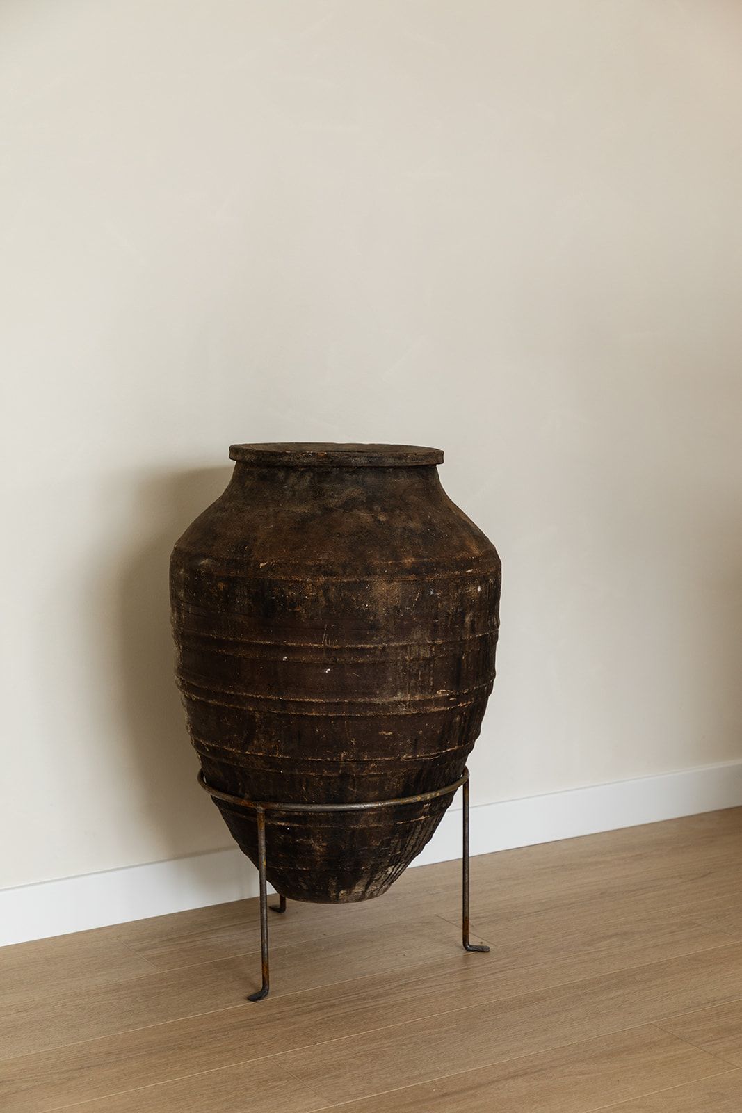 An antique earthenware storage vessel on metal stand against a white wall with wooden flooring.