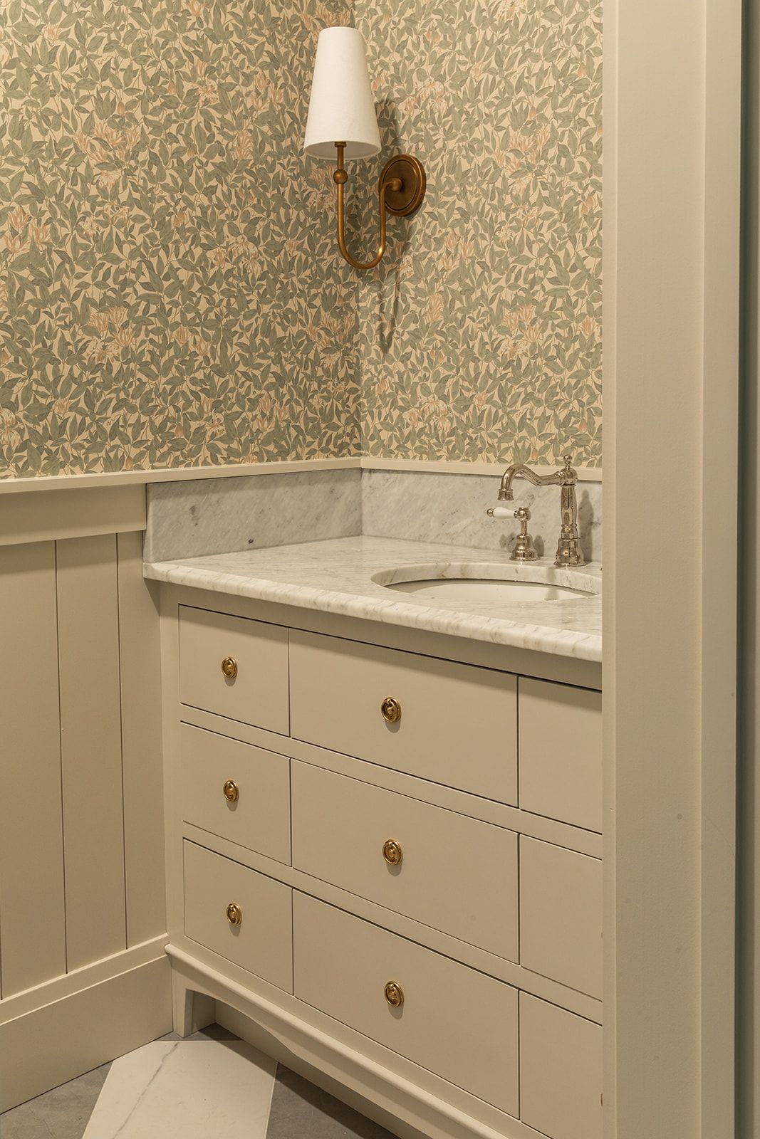 Elegant bathroom vanity with marble countertop, brass hardware, white drawers, and floral wallpaper with a gold wall sconce.