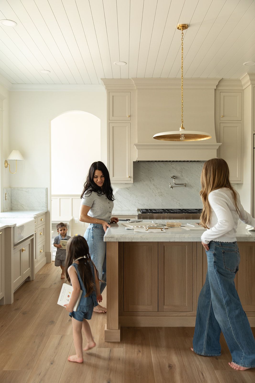 Woman and children gather around a kitchen island in a bright, modern kitchen with cream cabinets and wood floors.