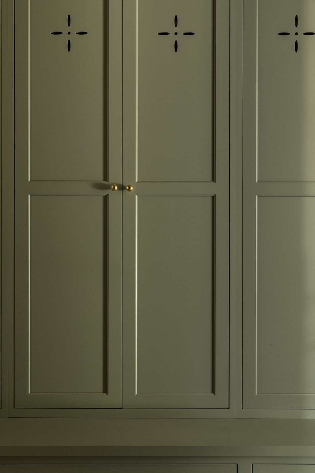 Sage green built-in wardrobe with decorative cross cutouts and brass hardware in a classic paneled cabinet design.
