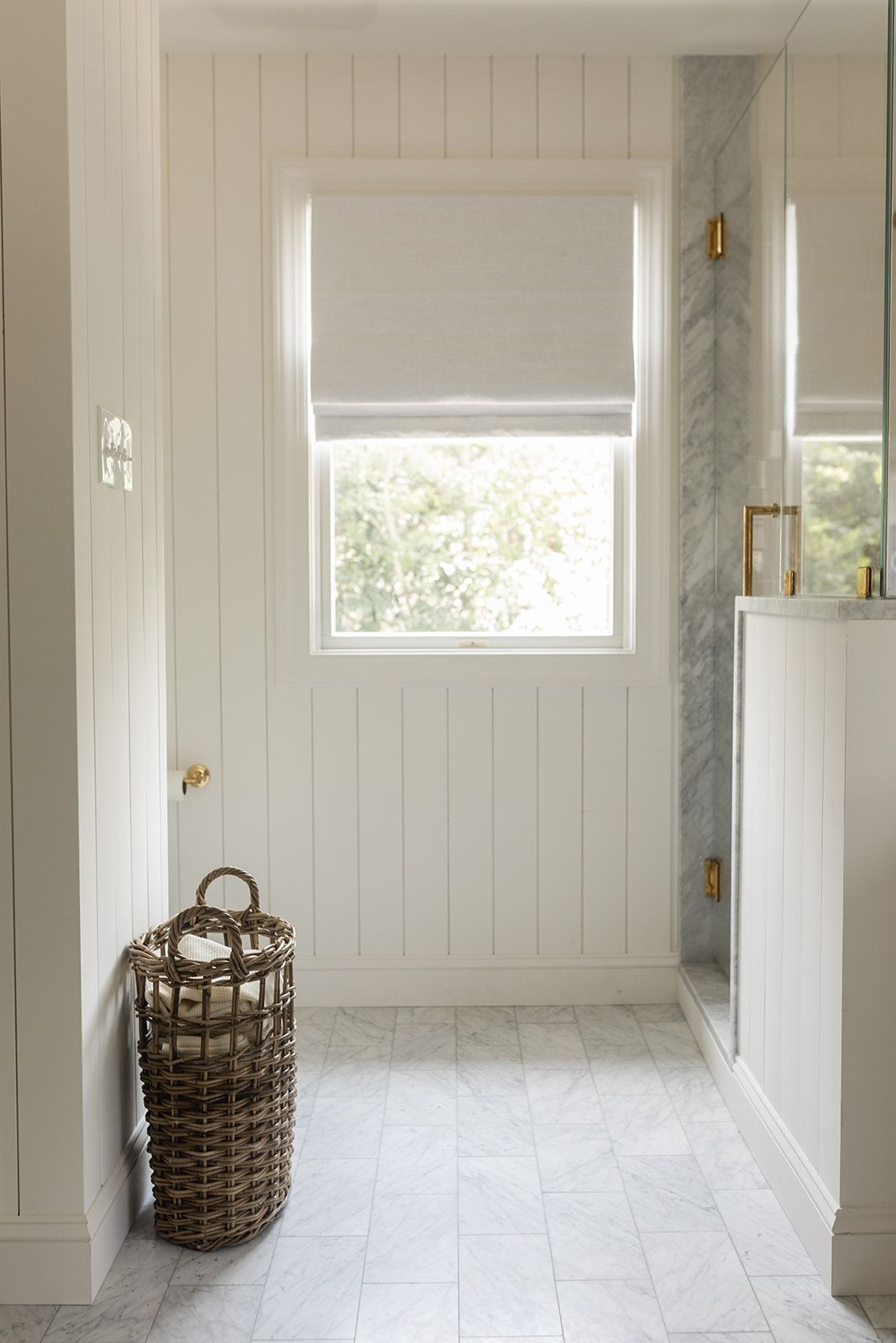 Bright white shiplap bathroom with marble tile floors, wicker basket, Roman shade window, and gold hardware accents.