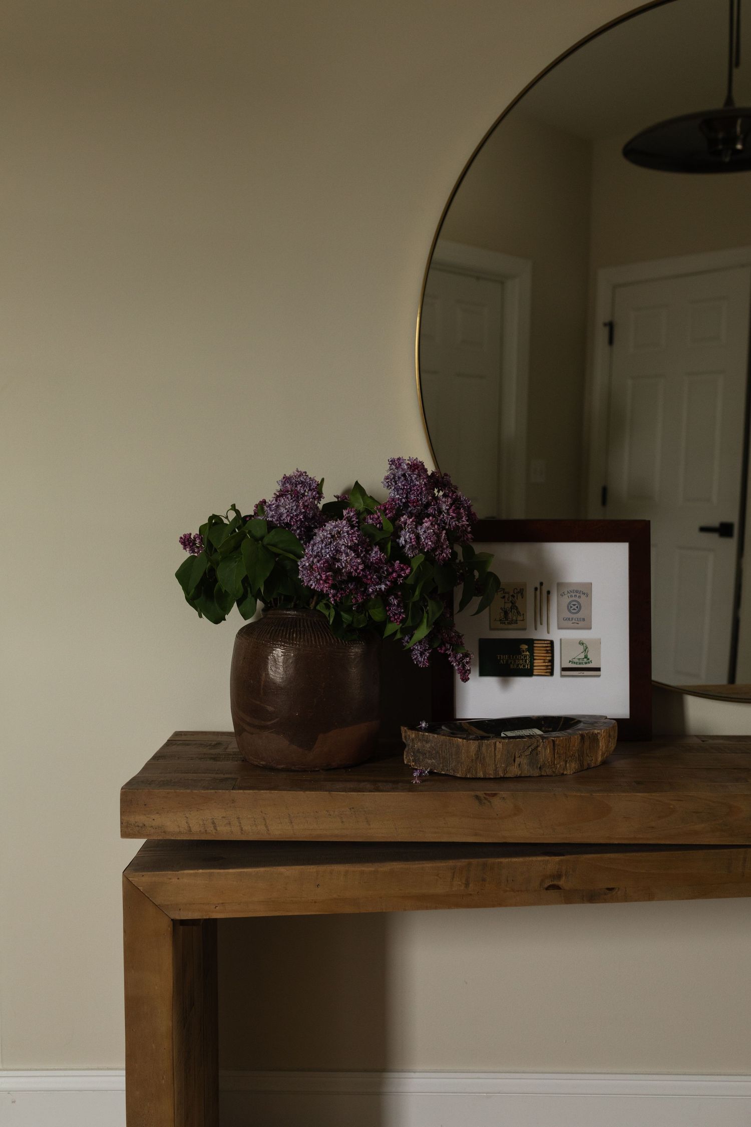 A wooden shelf mounted on a cream-colored wall holds a decorative brown pot filled with purple lilacs.