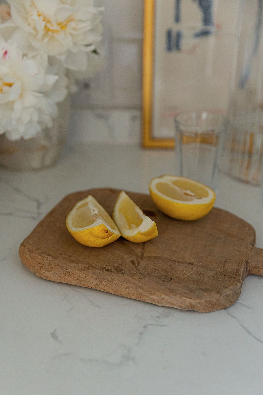 Fresh cut lemon halves on rustic wooden cutting board with white flowers and glassware in soft natural lighting.