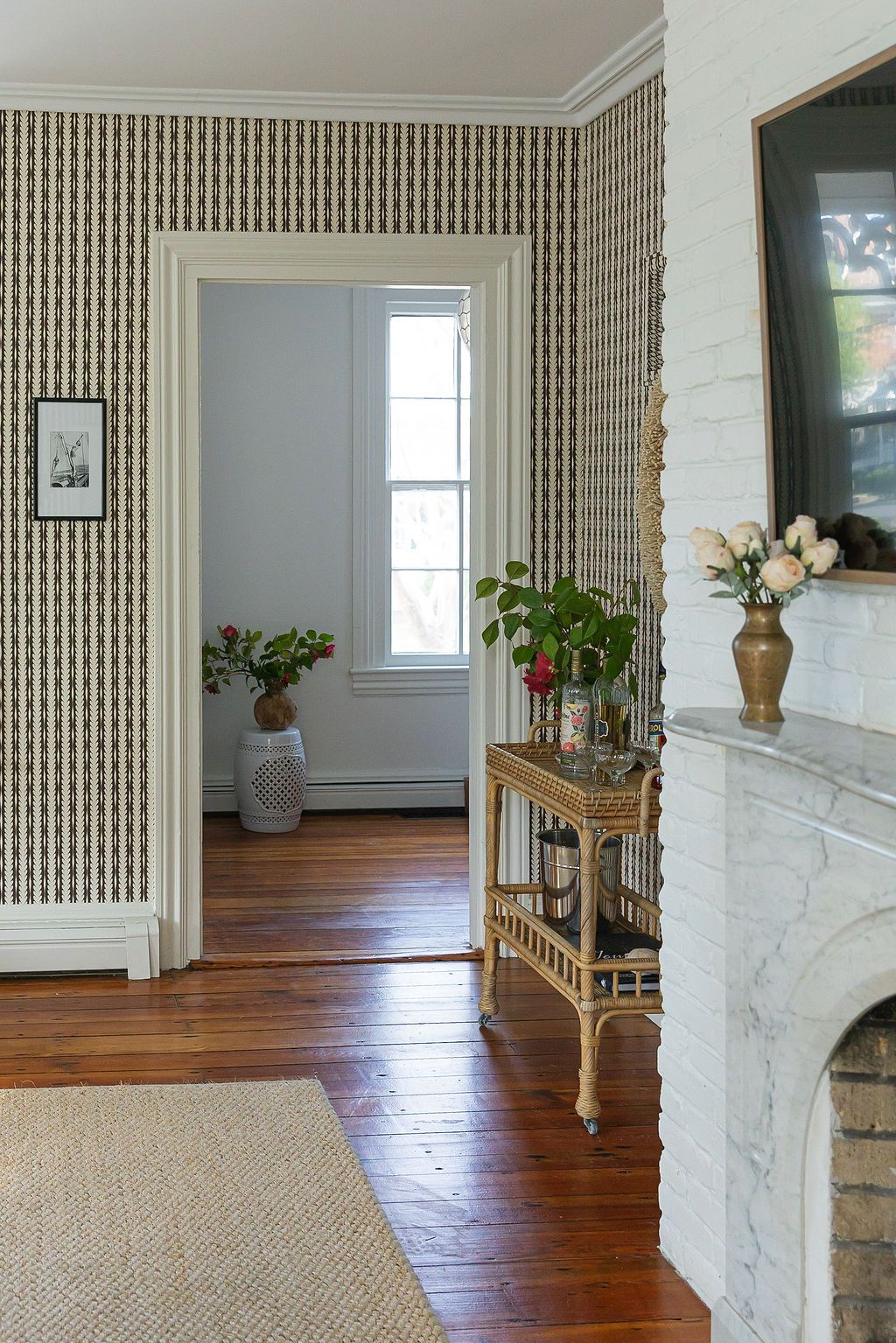 Hallway with striped wallpaper, white brick fireplace, and hardwood floors leads to bright doorway with indoor plants.