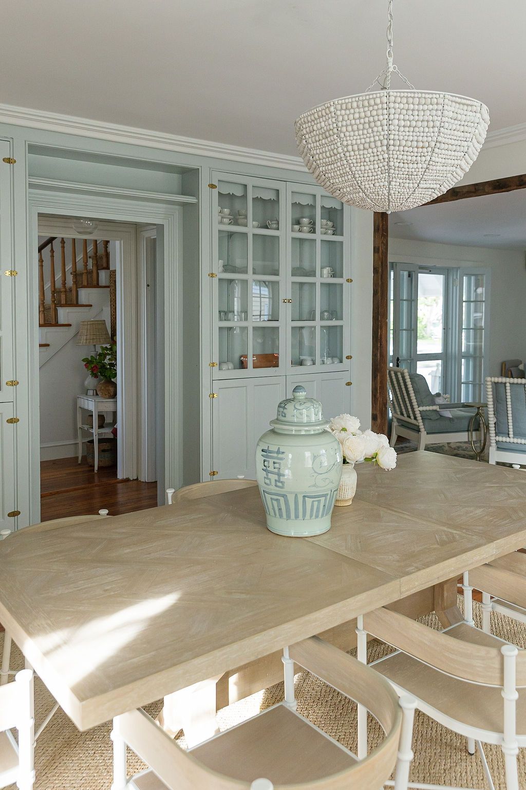 Coastal dining room with beaded chandelier, light blue cabinets, and a bleached wood table catching sunlight.