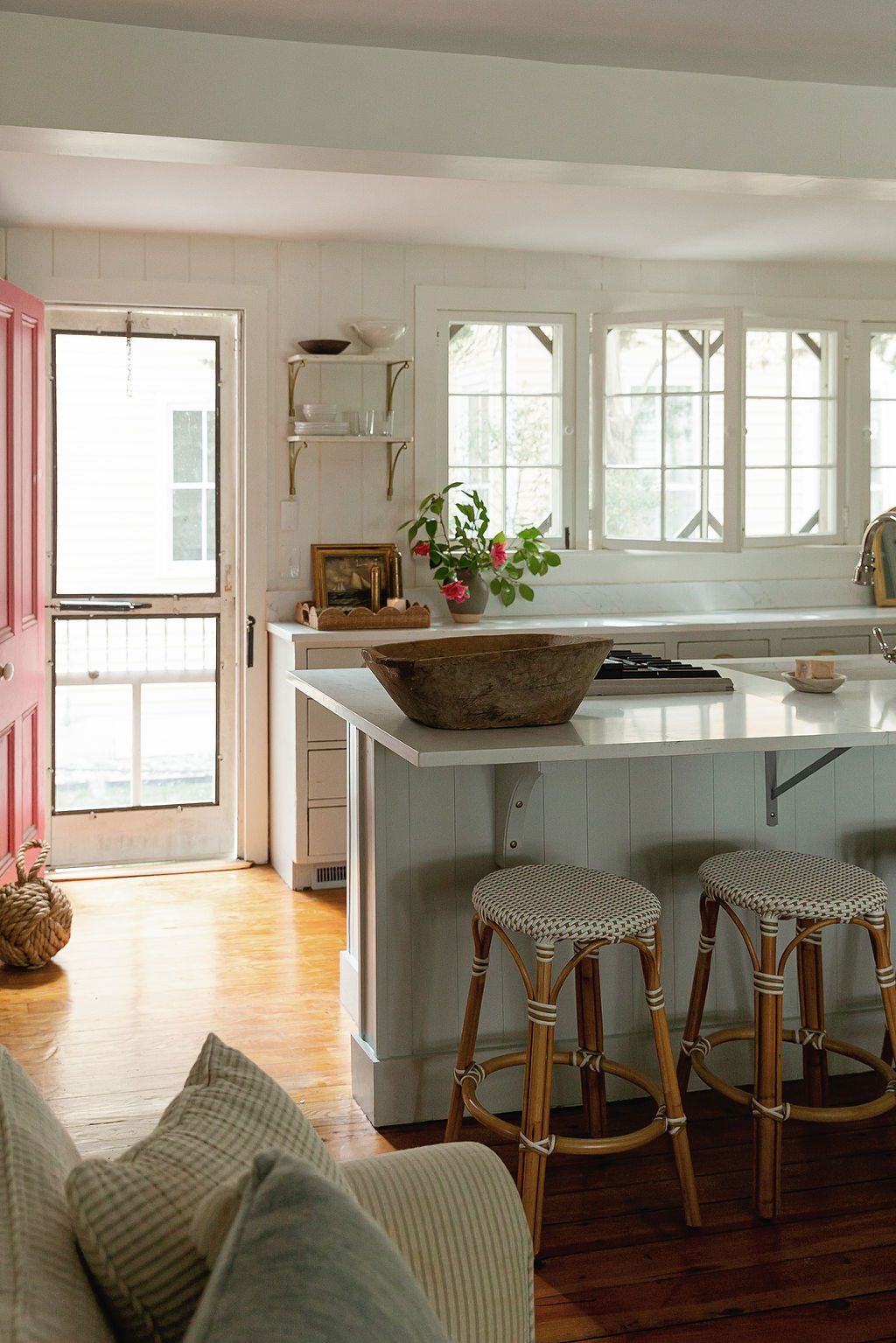 A cozy kitchen space with rattan bar stools, white windows, a wooden bowl centerpiece, and natural lighting.