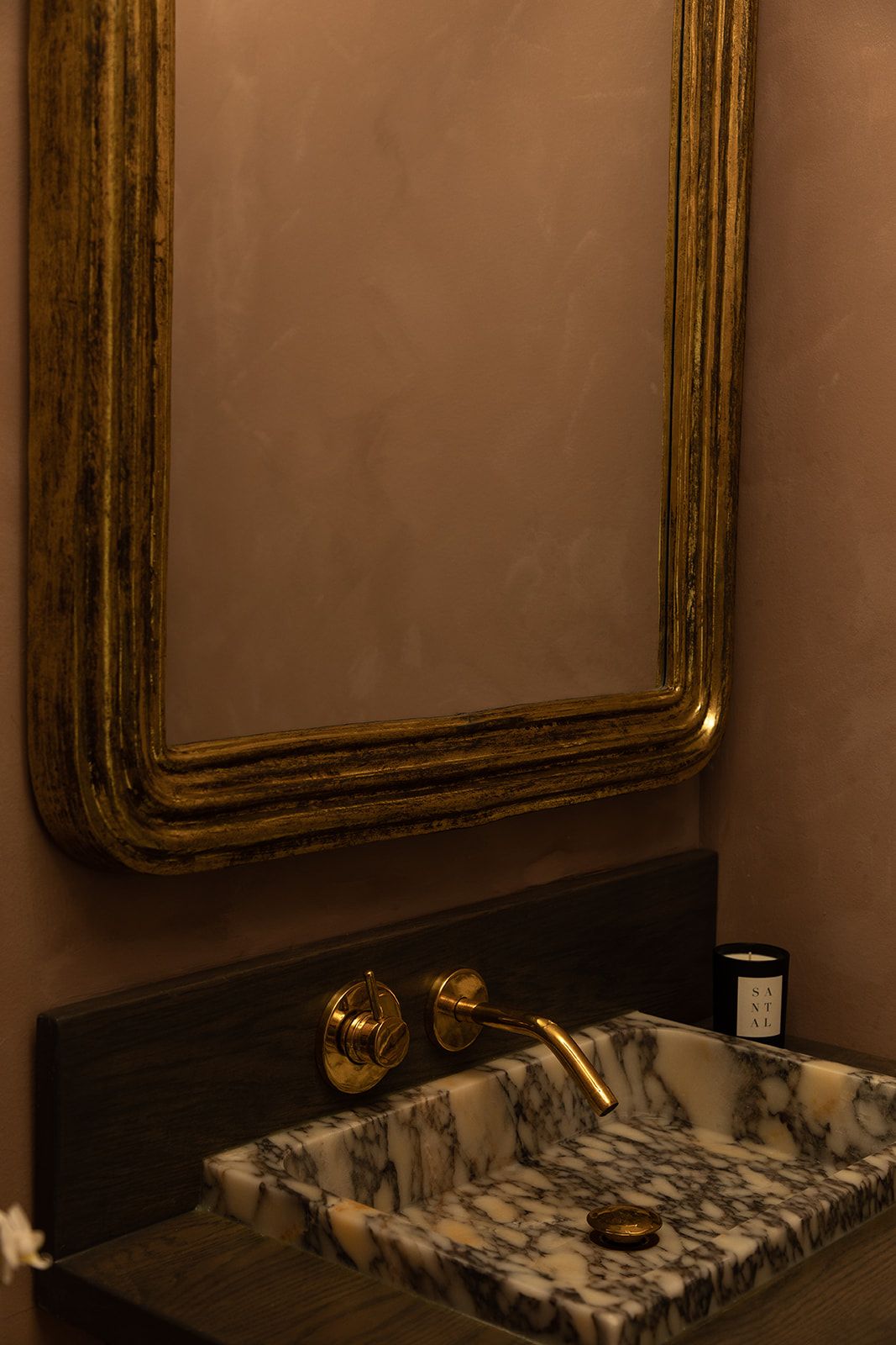 Vintage gold-framed mirror hangs above a marble vanity with brass fixtures in a moody bathroom setting.