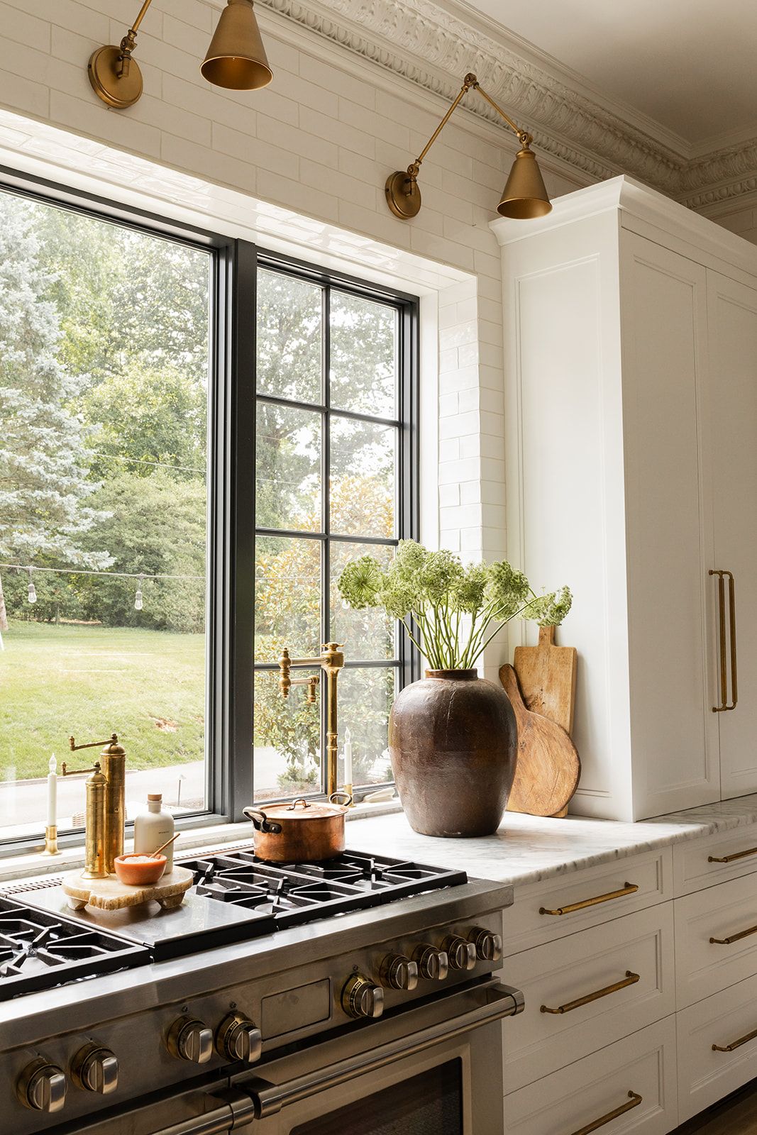 Bright kitchen window with brass sconces, gas range, and terra cotta vase with greenery.