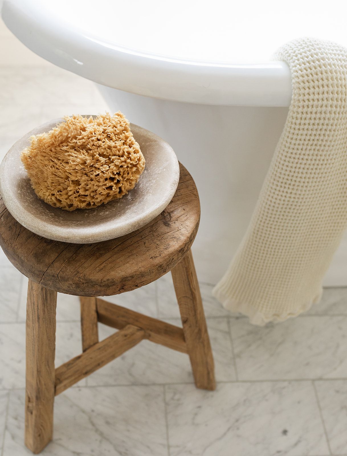 Rustic wooden stool with natural sea sponge on marble dish beside white freestanding bathtub with waffle towel.
