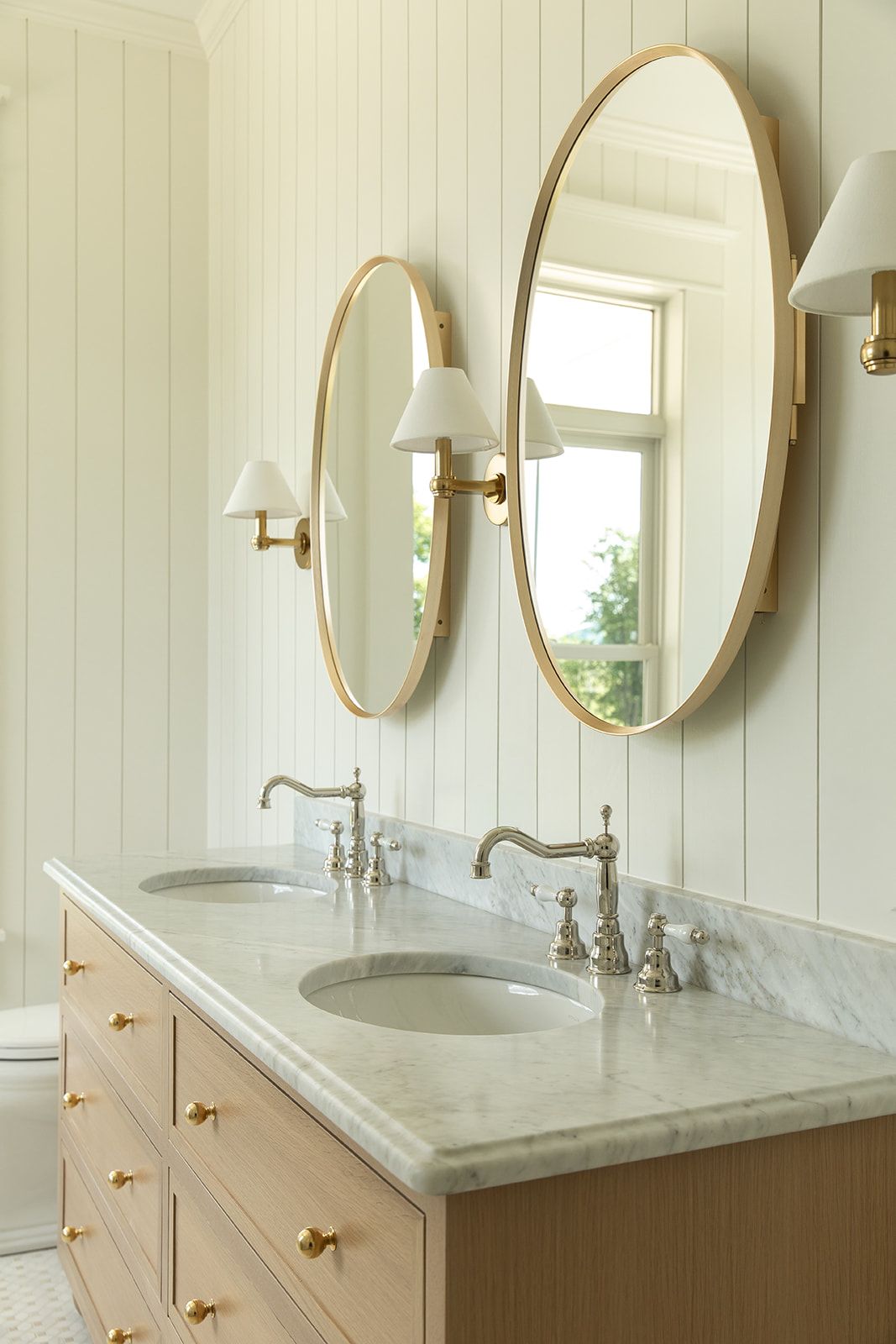 Elegant double bathroom vanity with marble countertop, wood cabinets, oval gold mirrors, and brass wall sconces on white paneled walls.