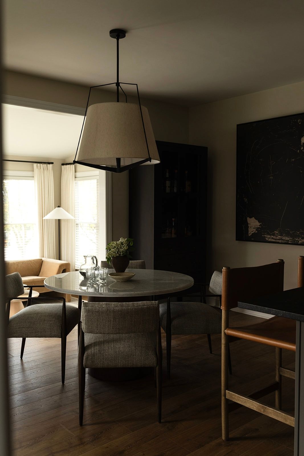 A moody dining room interior with round table, upholstered chairs, and pendant light fixture in dark atmospheric lighting.