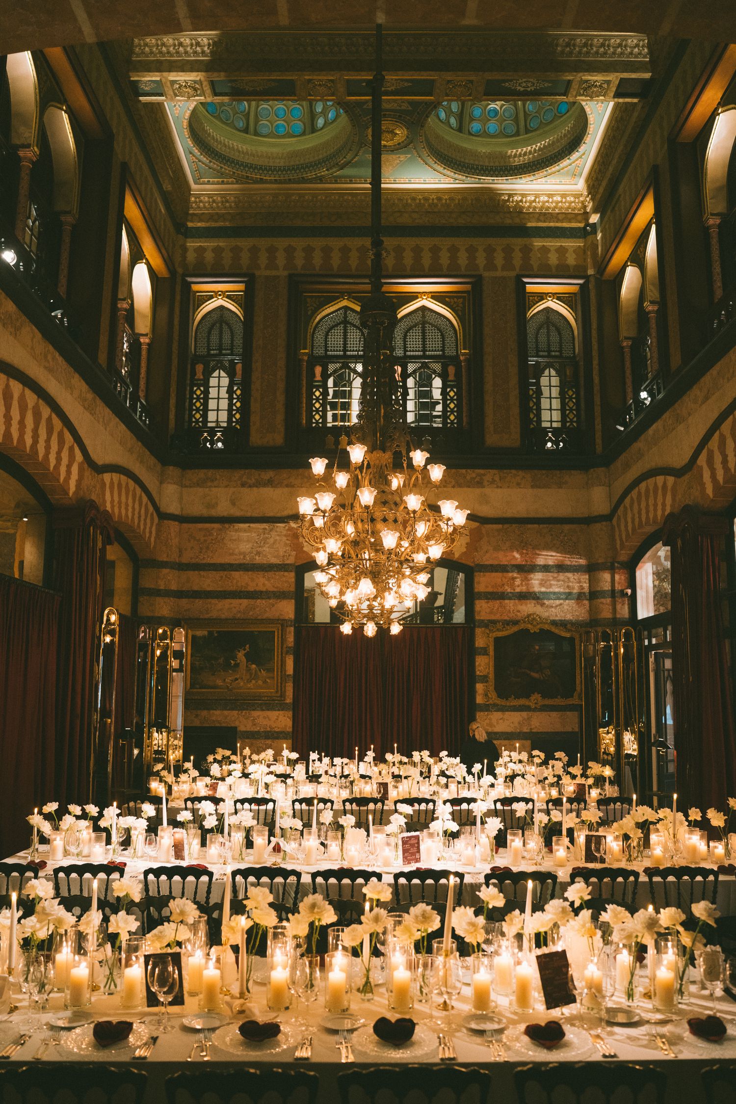 Elegant wedding reception tables with glowing candles and white decor in a grand historic ballroom with ornate chandeliers.