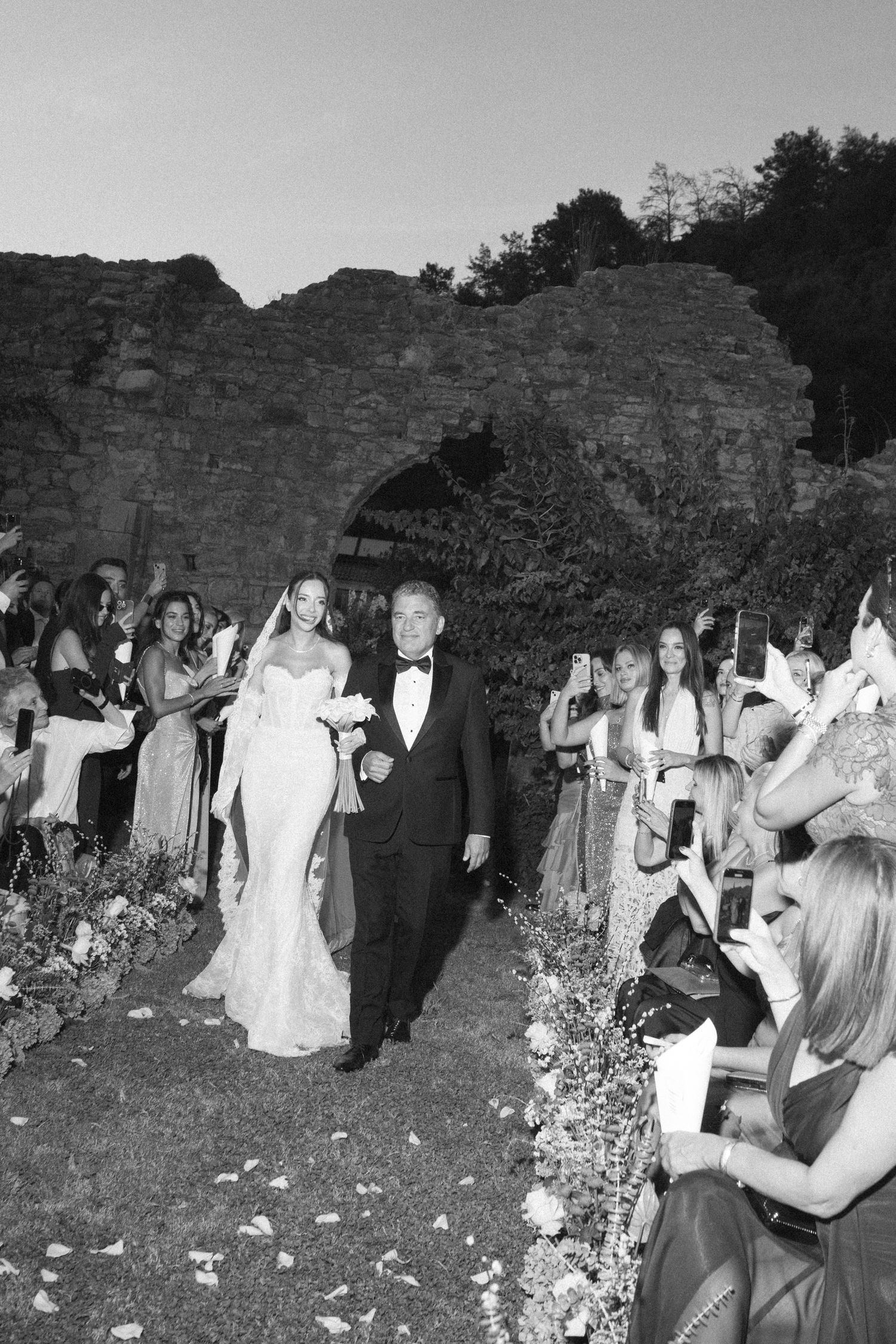 A black and white photograph shows people in white robes gathered along a stone wall at an outdoor event.