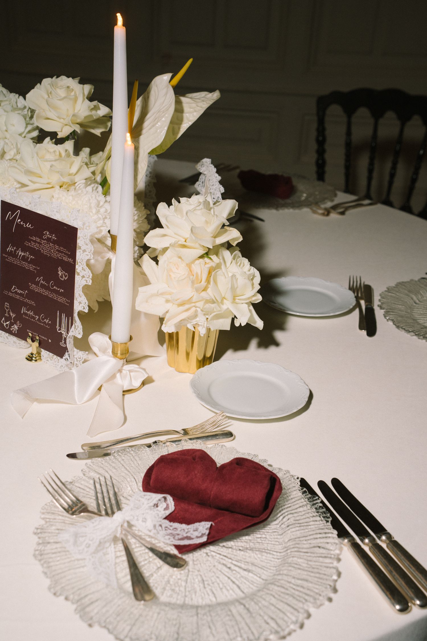 Elegant table setting with white candle, flowers, burgundy napkin, and vintage silverware on white tablecloth.