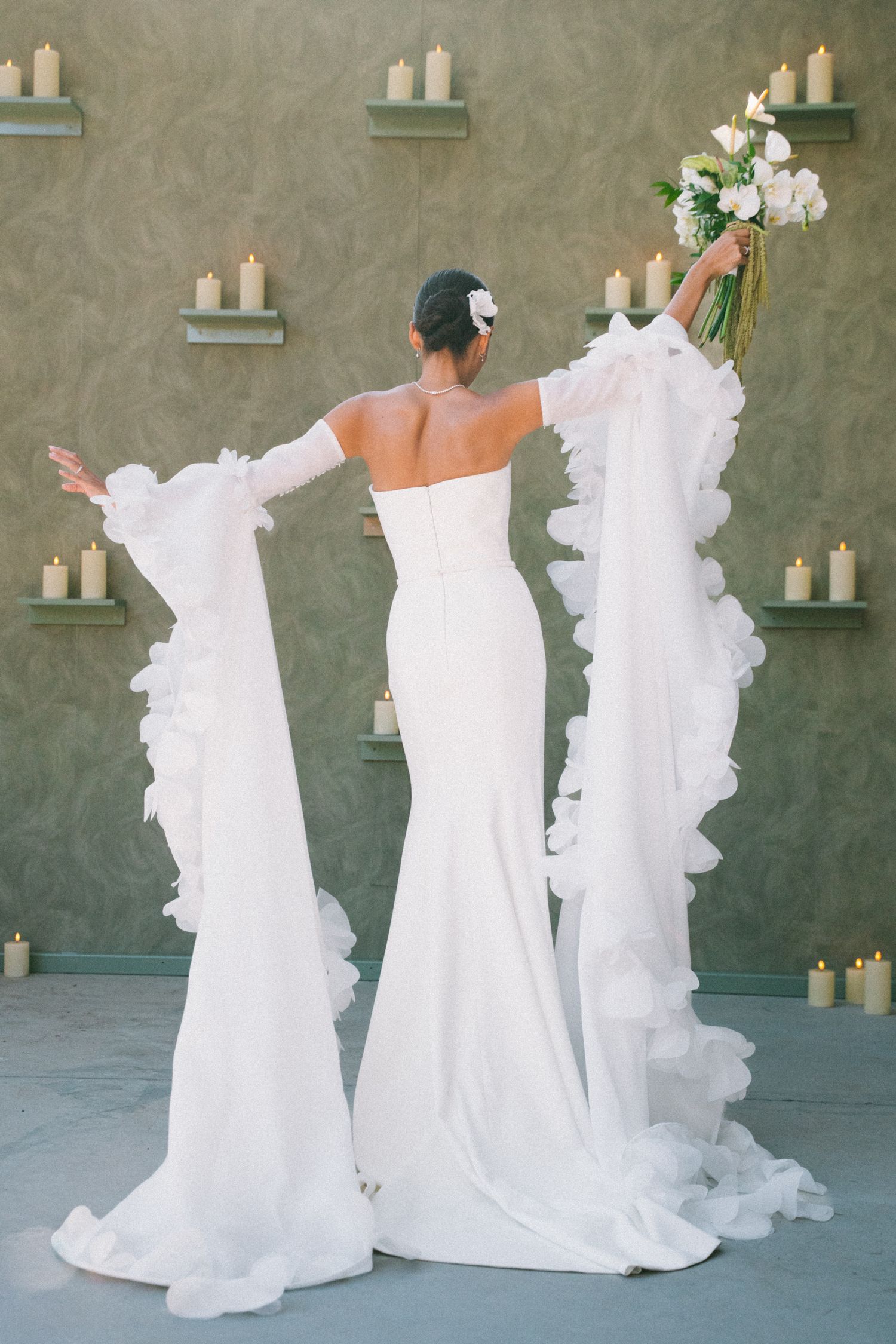 Elegant white wedding dress with dramatic ruffled train displayed against sage green wall with floating candle shelves.