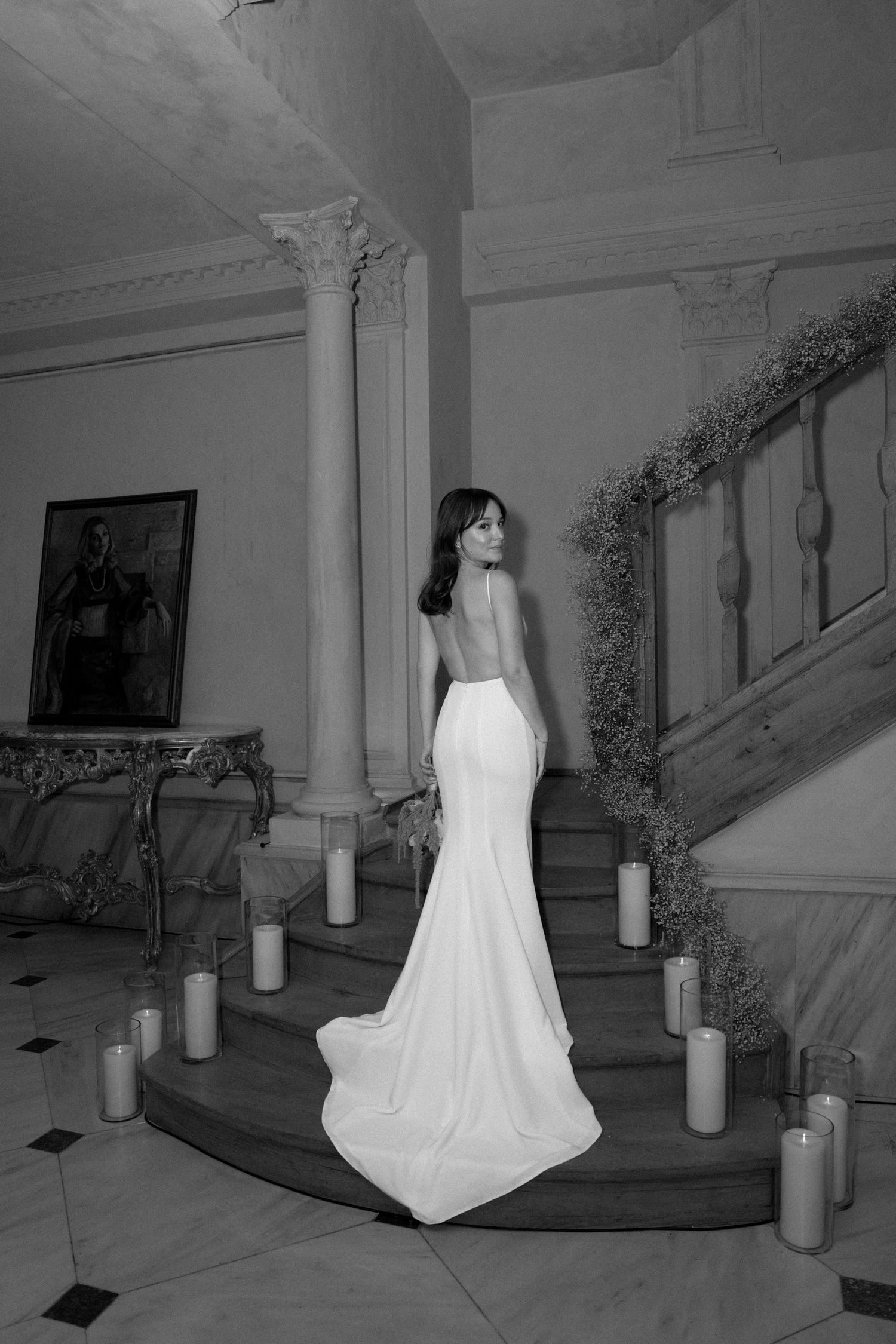 A bride in a backless white mermaid wedding gown stands on an elegant staircase surrounded by glowing candles.