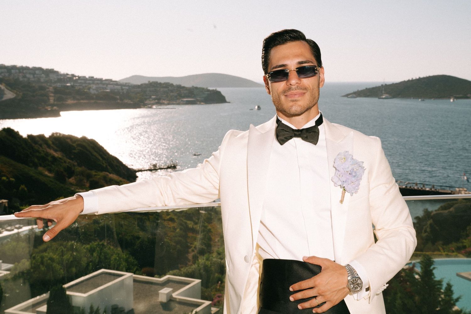 A person in a white tuxedo and sunglasses poses against a scenic coastal backdrop with water and hills in view.