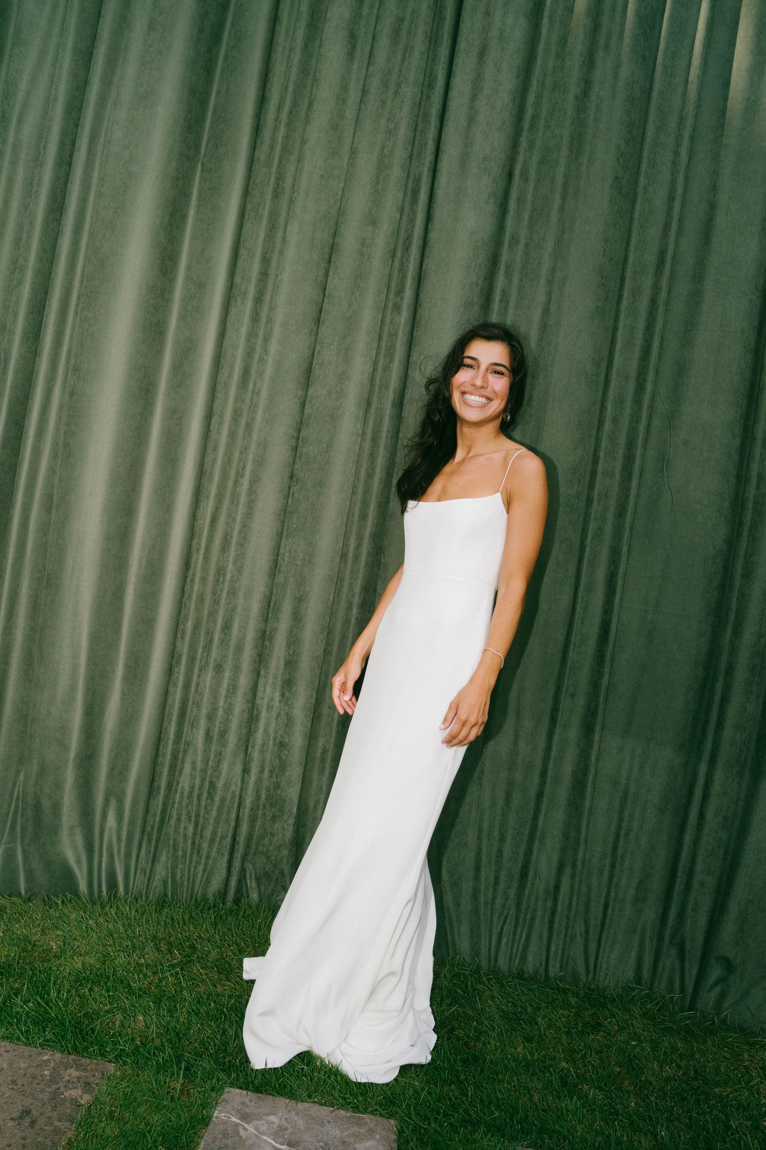 Smiling woman in a white spaghetti strap gown standing on grass in front of a green velvet curtain backdrop.