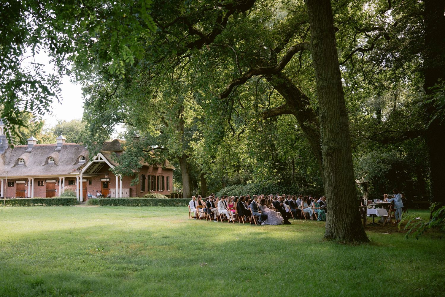 Garden wedding ceremony with guests gathered under large trees near a rustic brick building.