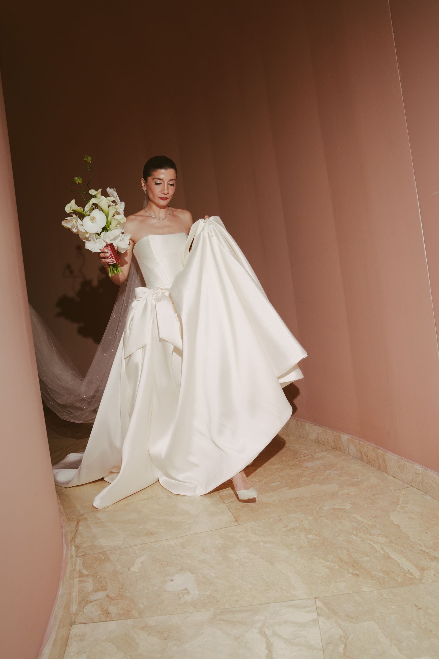 Bride in strapless white satin ballgown with bow belt holds floral bouquet in pink marble hallway.