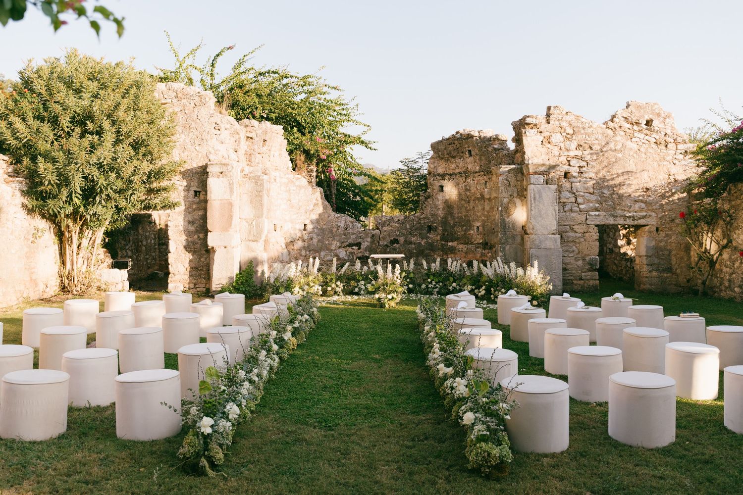 White cylindrical seating arrangements line a grassy outdoor wedding aisle decorated with floral arrangements at sunset.