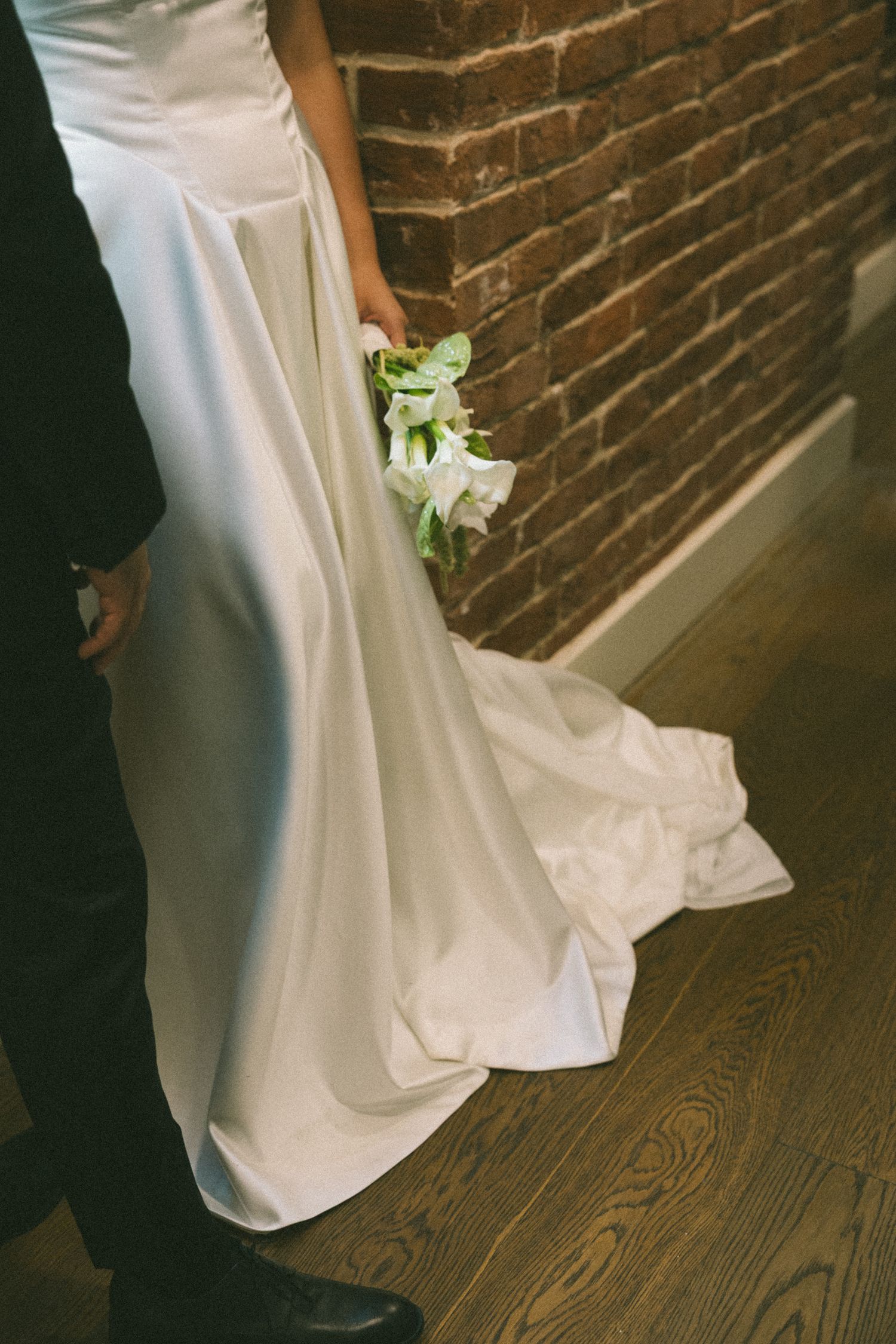A partial view of a white wedding dress train and small bouquet against a brick wall and wooden floor.