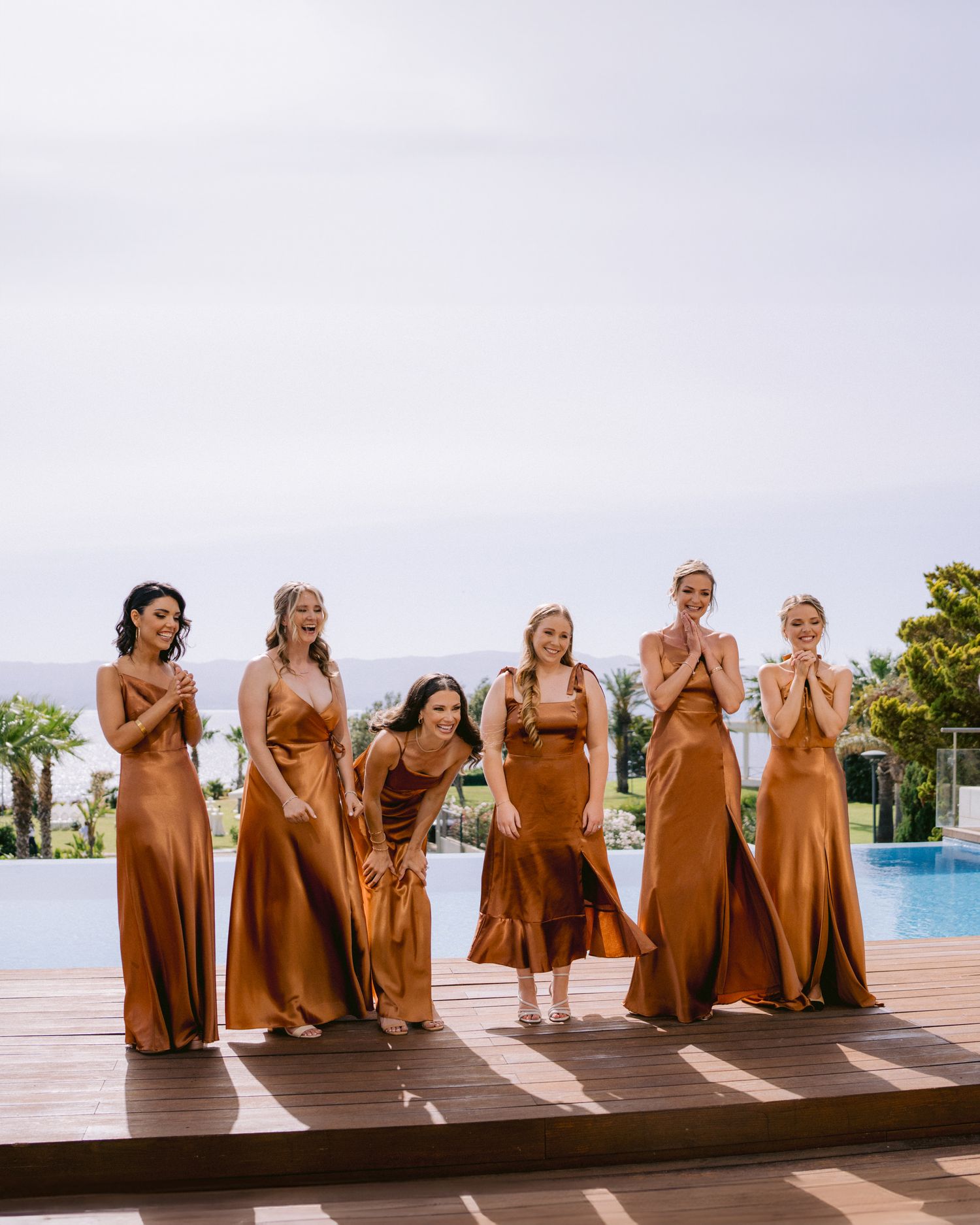 Bridesmaids in matching copper-colored dresses pose by a poolside setting.