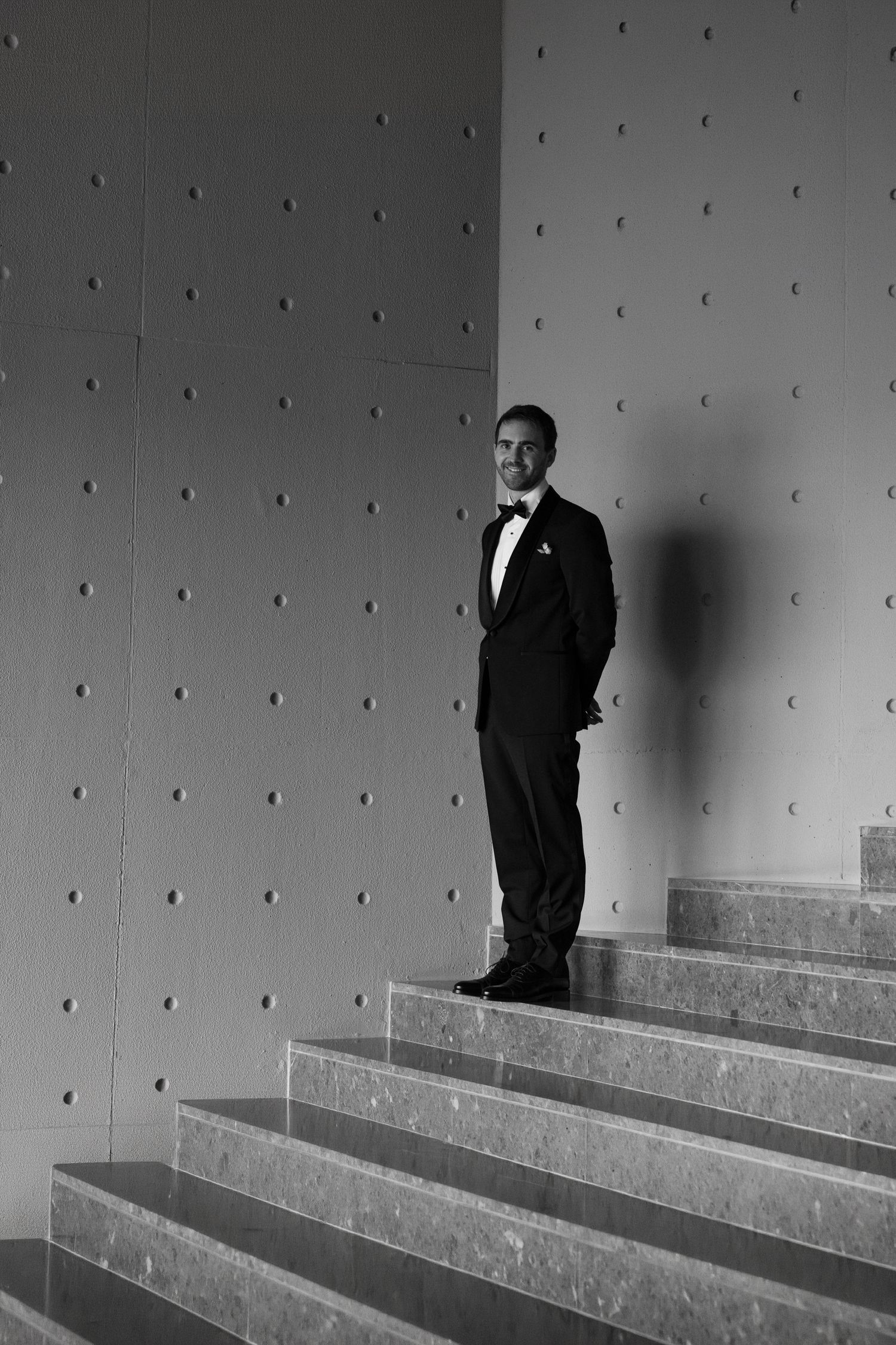 Elegant man in tuxedo standing on marble stairs against a dotted wall in a black and white photo.