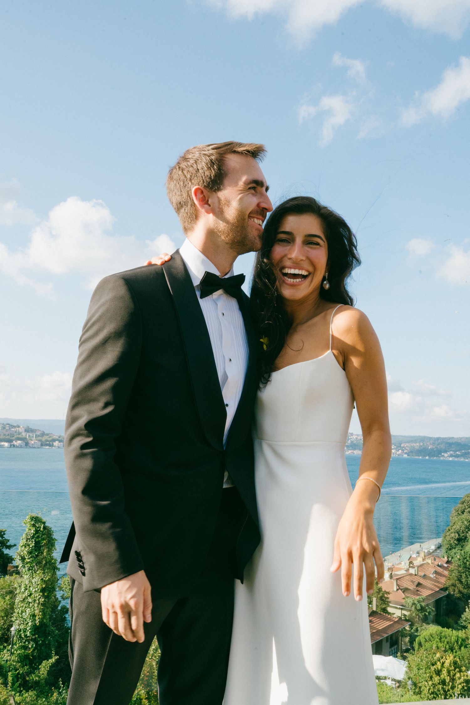 A joyful couple in wedding attire laughing together with a scenic waterfront view and blue sky behind them.