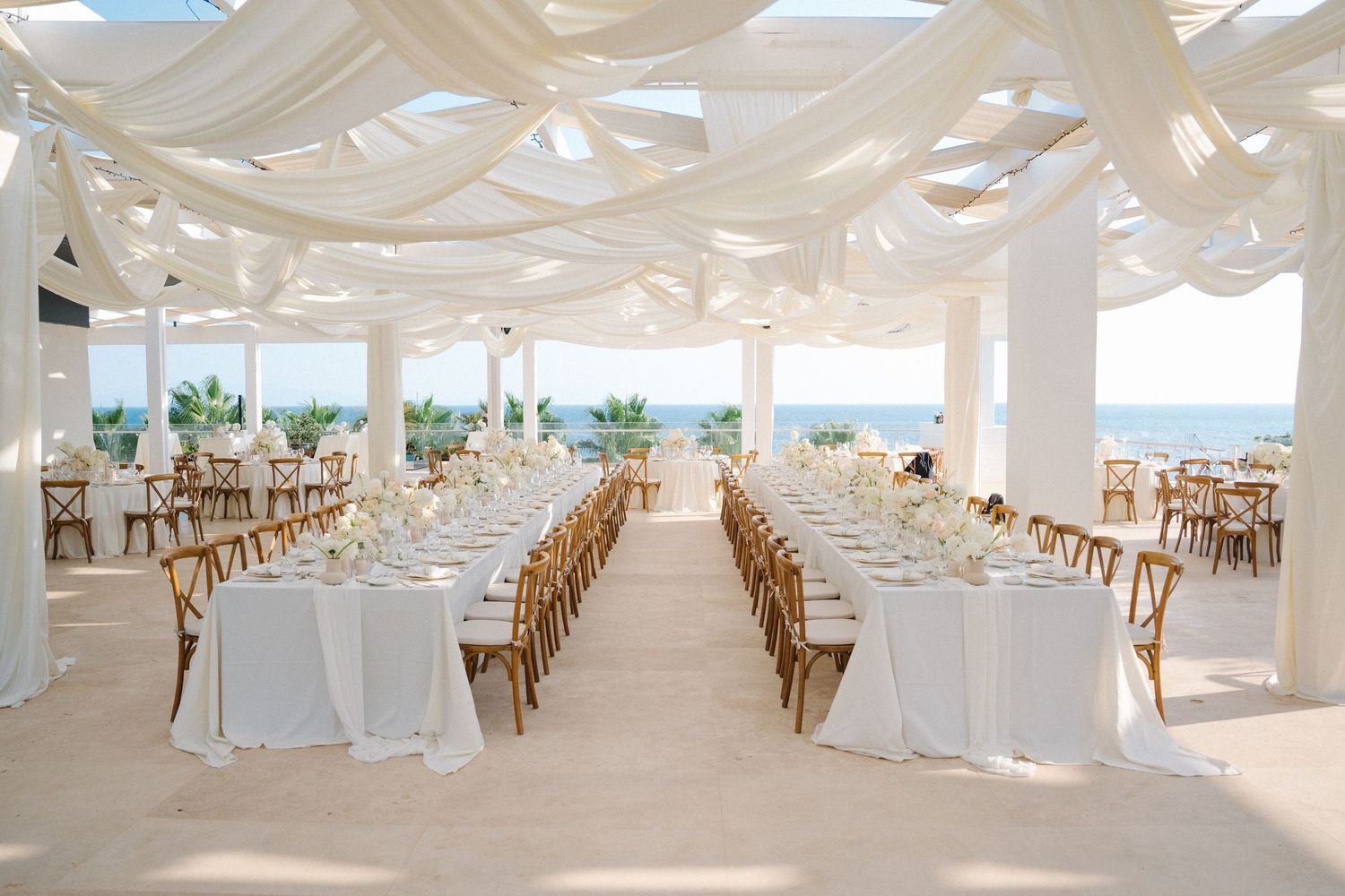 Elegant white wedding reception venue with draped fabric ceilings, wooden chairs and ocean views through large windows.