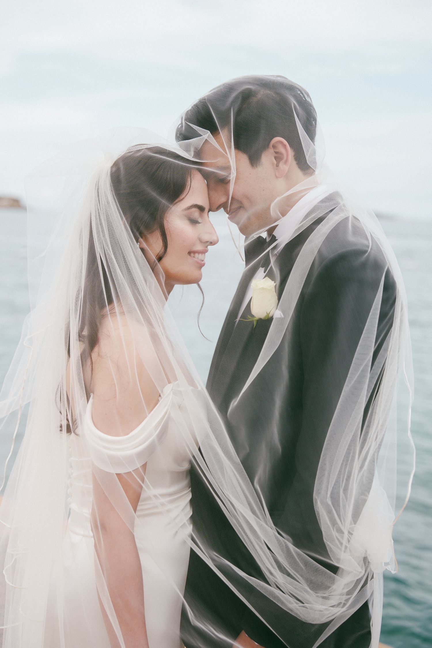 Bride and groom sharing an intimate moment under a flowing white veil by the ocean on their wedding day.