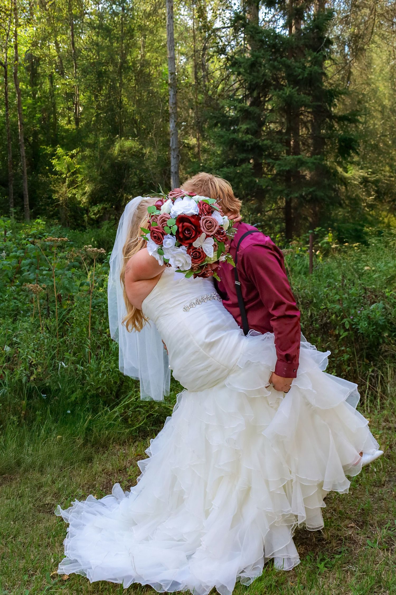 A couple in wedding attire dips dramatically in a romantic pose against a lush forest backdrop.