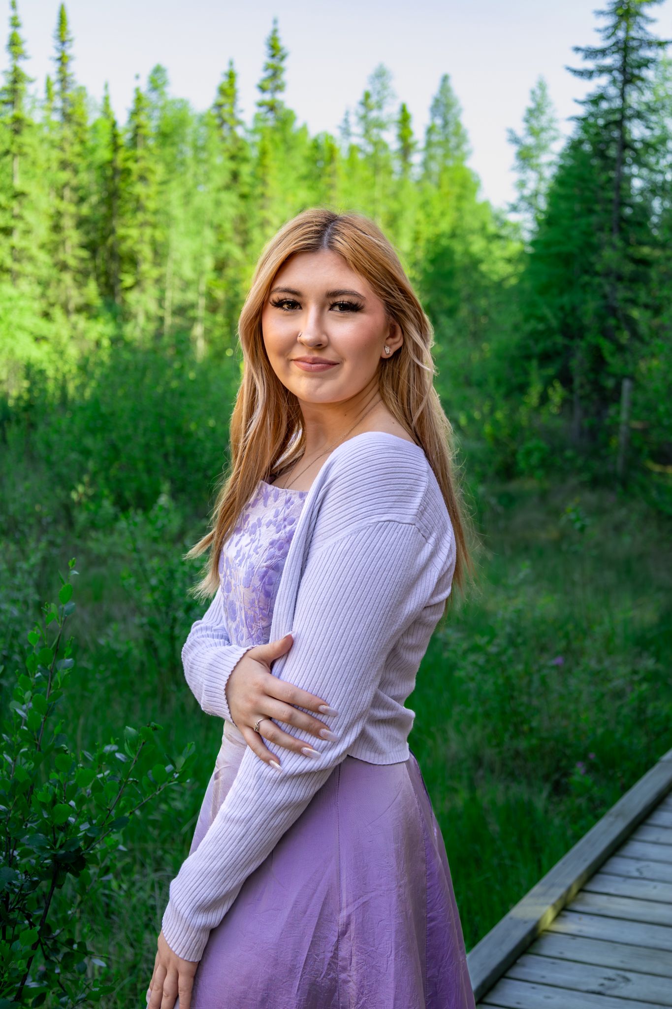 A person in a lavender dress poses on a wooden boardwalk surrounded by evergreen trees.